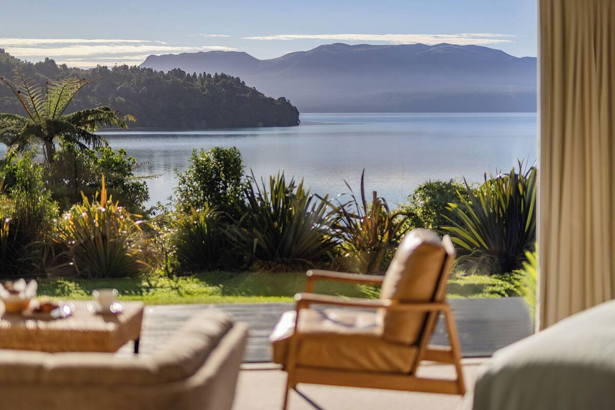 A room with a view of a lake and mountains. There is a chair and a table with a glass of water.