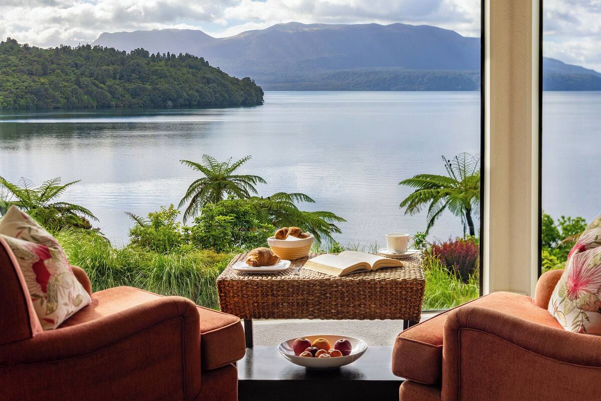 A room with a view of a lake and mountains, furnished with two sofas and a table with a bowl of fruit and a book.
