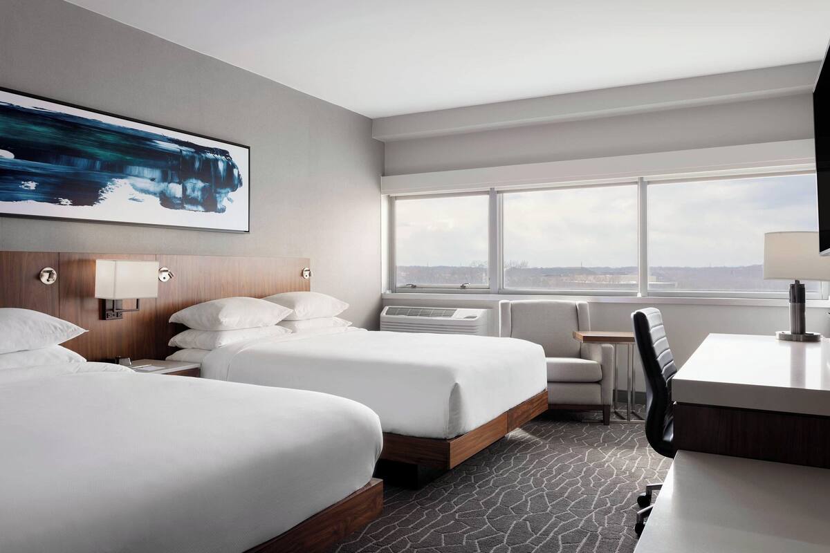 A hotel room with two beds, a desk, a chair, a window with a city view, and a painting on the wall.