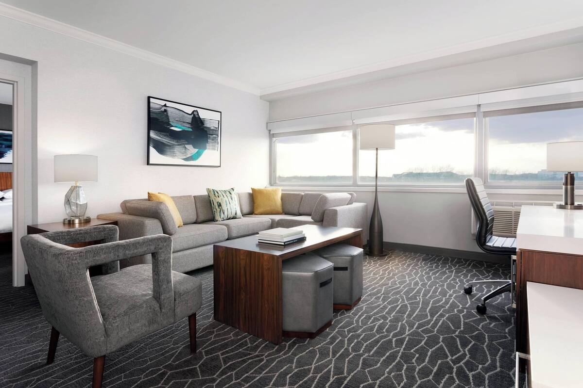 A modern hotel room with a gray sofa set, a wooden coffee table, a desk, and a large window offering a view of the city.