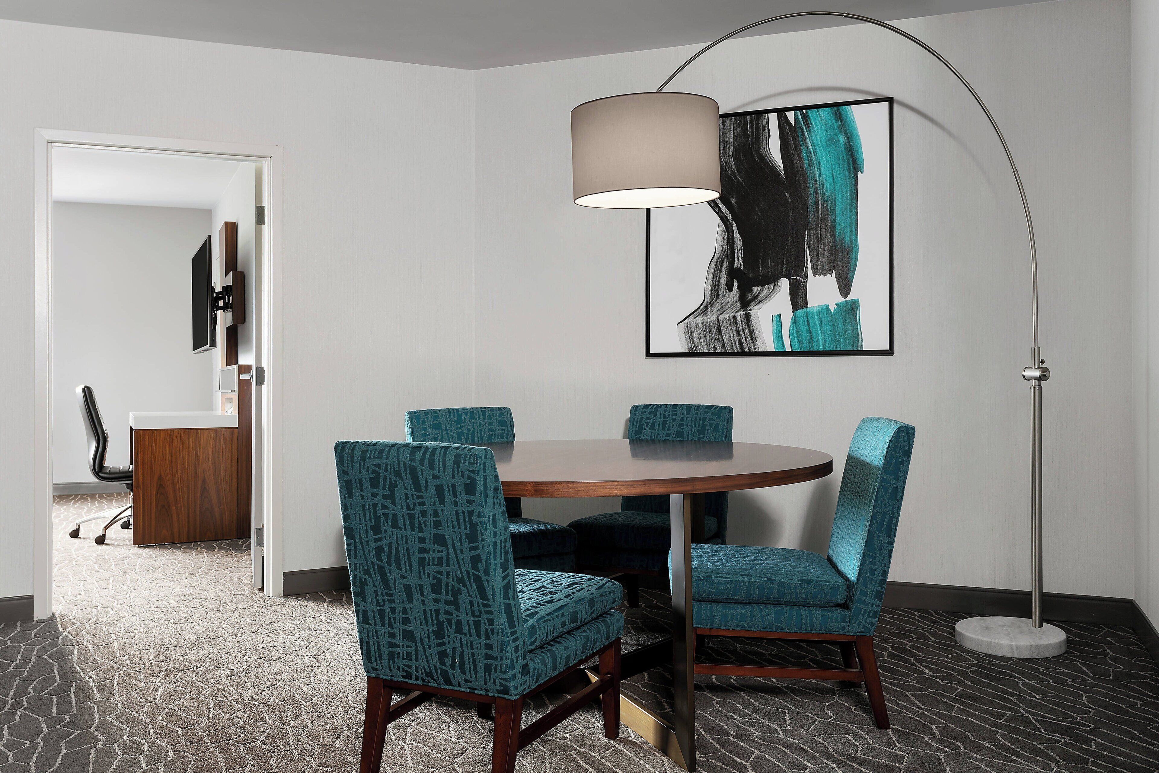 A conference room with a round table, four teal chairs, a modern floor lamp, and a large abstract painting on the wall.