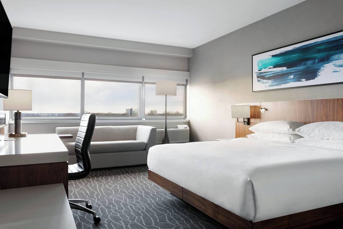 A modern hotel room with a large bed, a desk, a chair, and a view of the city skyline through large windows.