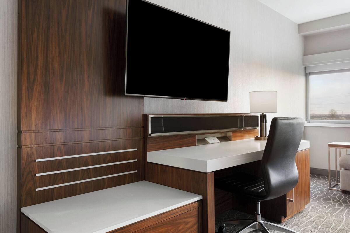 A modern hotel room with a flat-screen TV mounted on the wall, a wooden desk with a white surface, a black office chair, and a small table with a lamp.