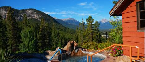 A hot tub with a mountain backdrop, surrounded by greenery and a wooden building.