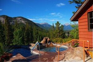 Un jacuzzi con montañas al fondo, rodeado de vegetación y un edificio de madera.