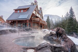 Un chalet à la façade en pierre, doté de balcons en bois et d’un espace dédié aux sources chaudes, où l’on peut voir de la vapeur s’élever.