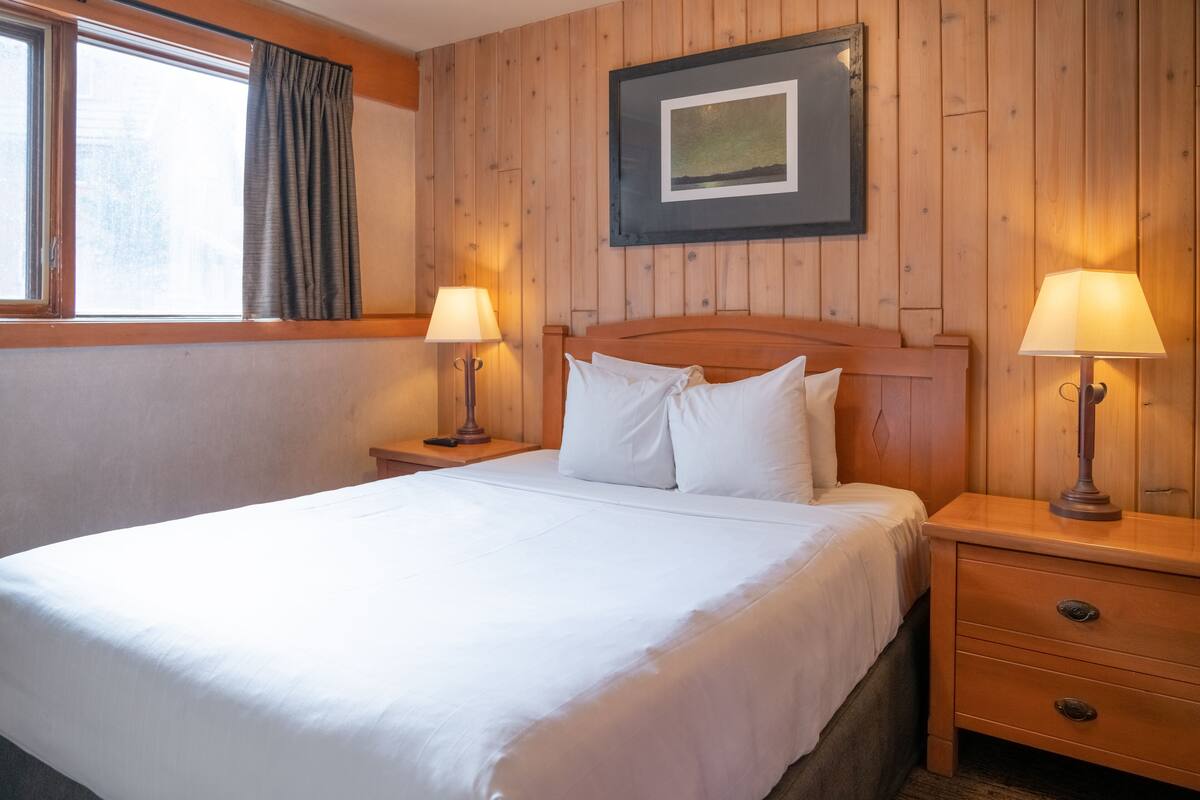 A neatly made bed in a room with wooden panel walls, two bedside tables with lamps, and a framed picture above the bed.