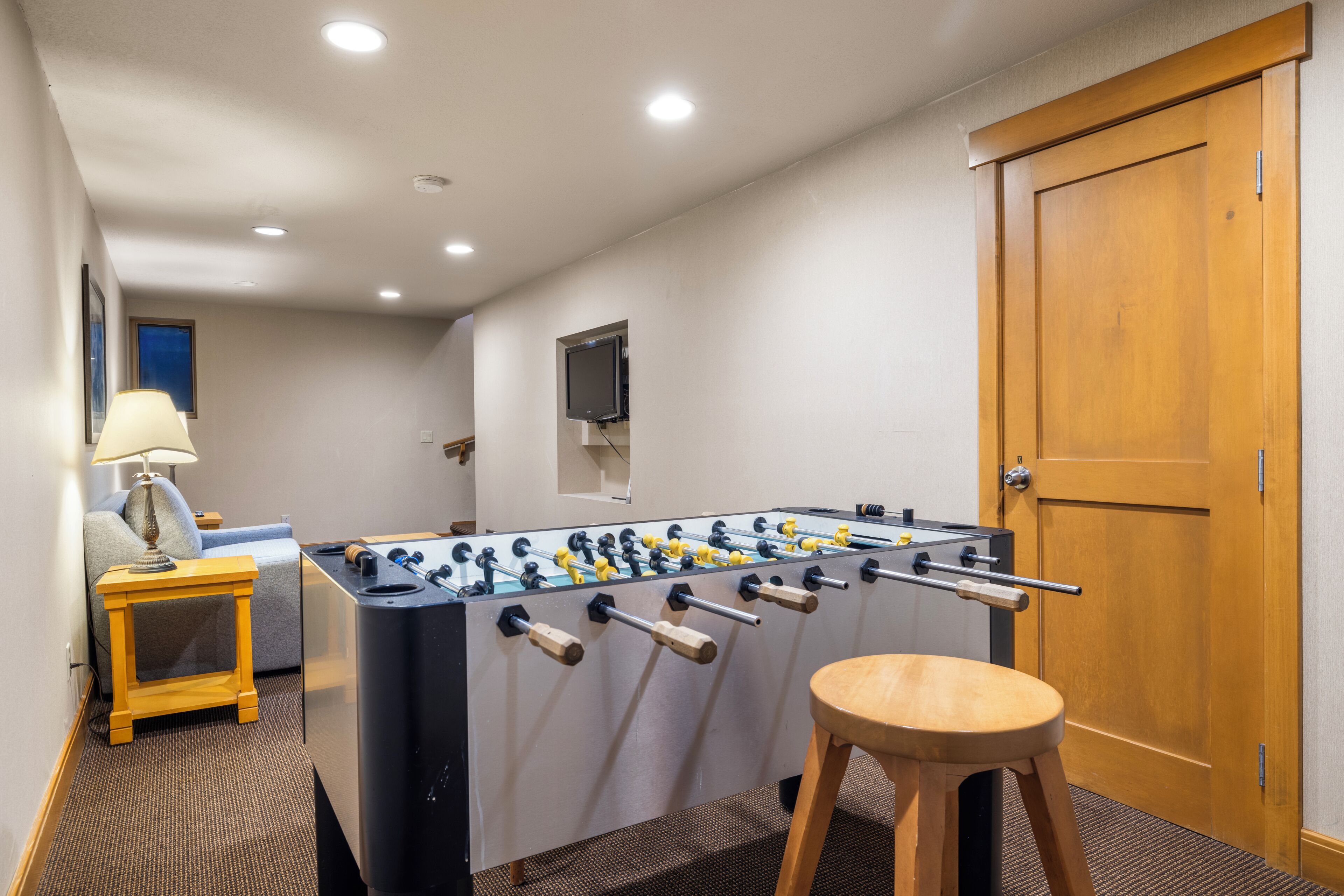 A room with a foosball table, a wooden stool, a lamp, and a door.