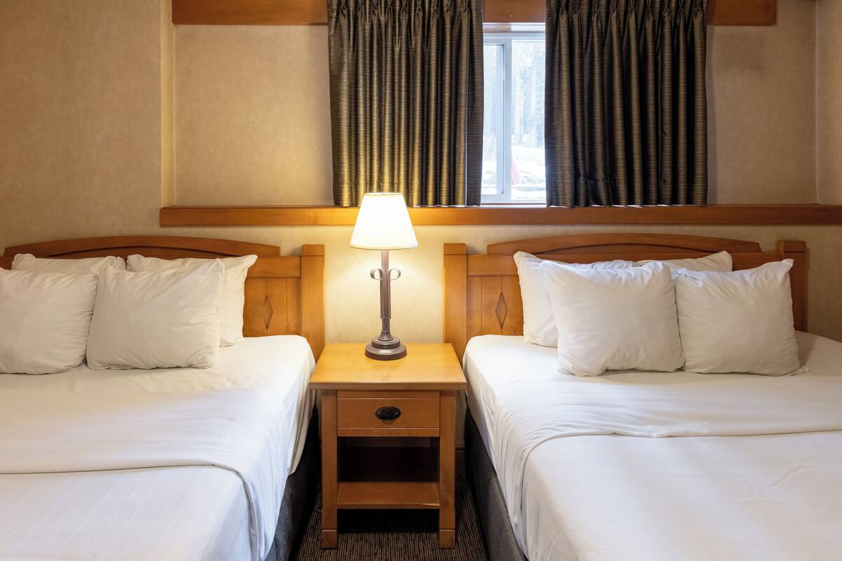 A hotel room with two beds, a nightstand with a lamp, and a window with curtains.