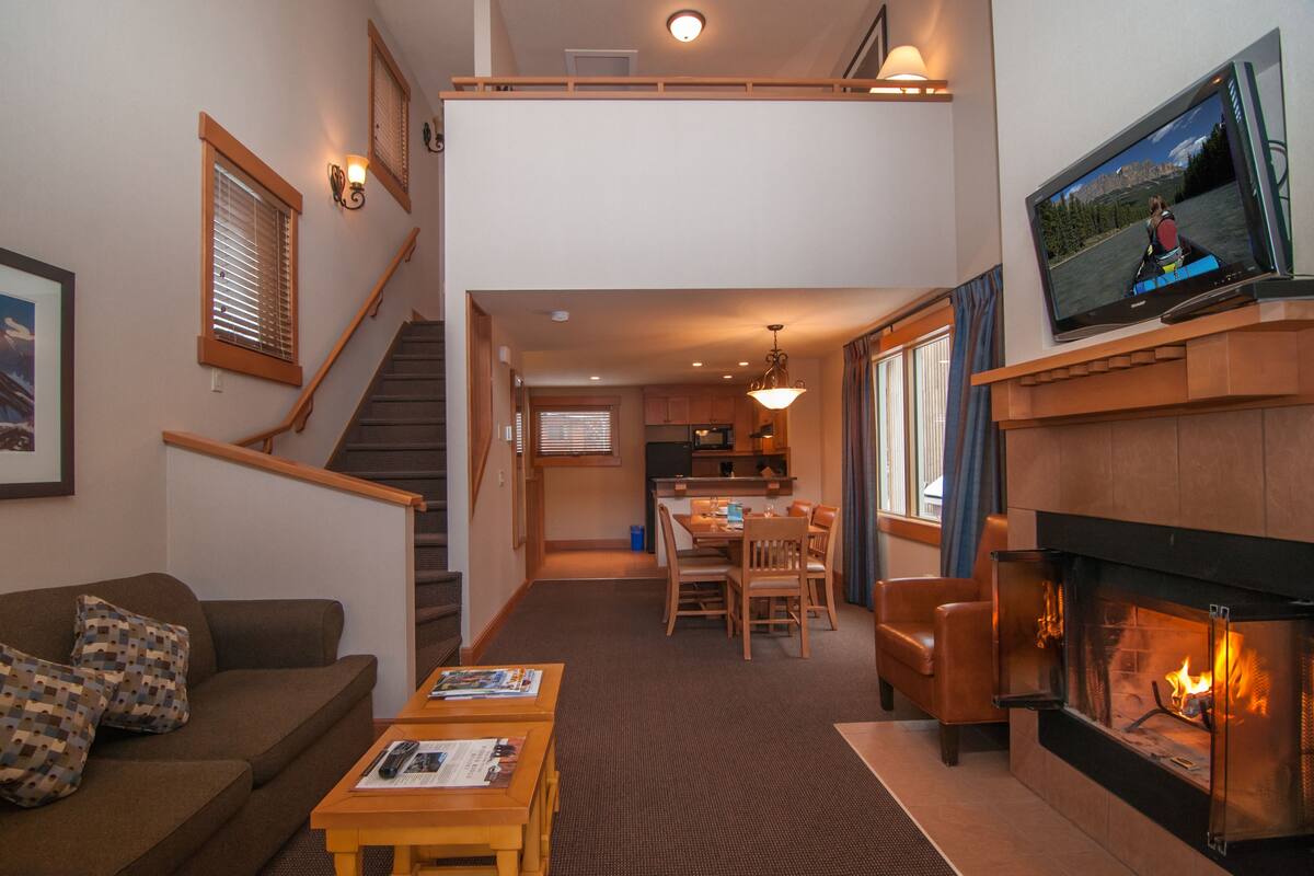 A living room with a fireplace, a television, a dining area, and a staircase leading to an upper level.