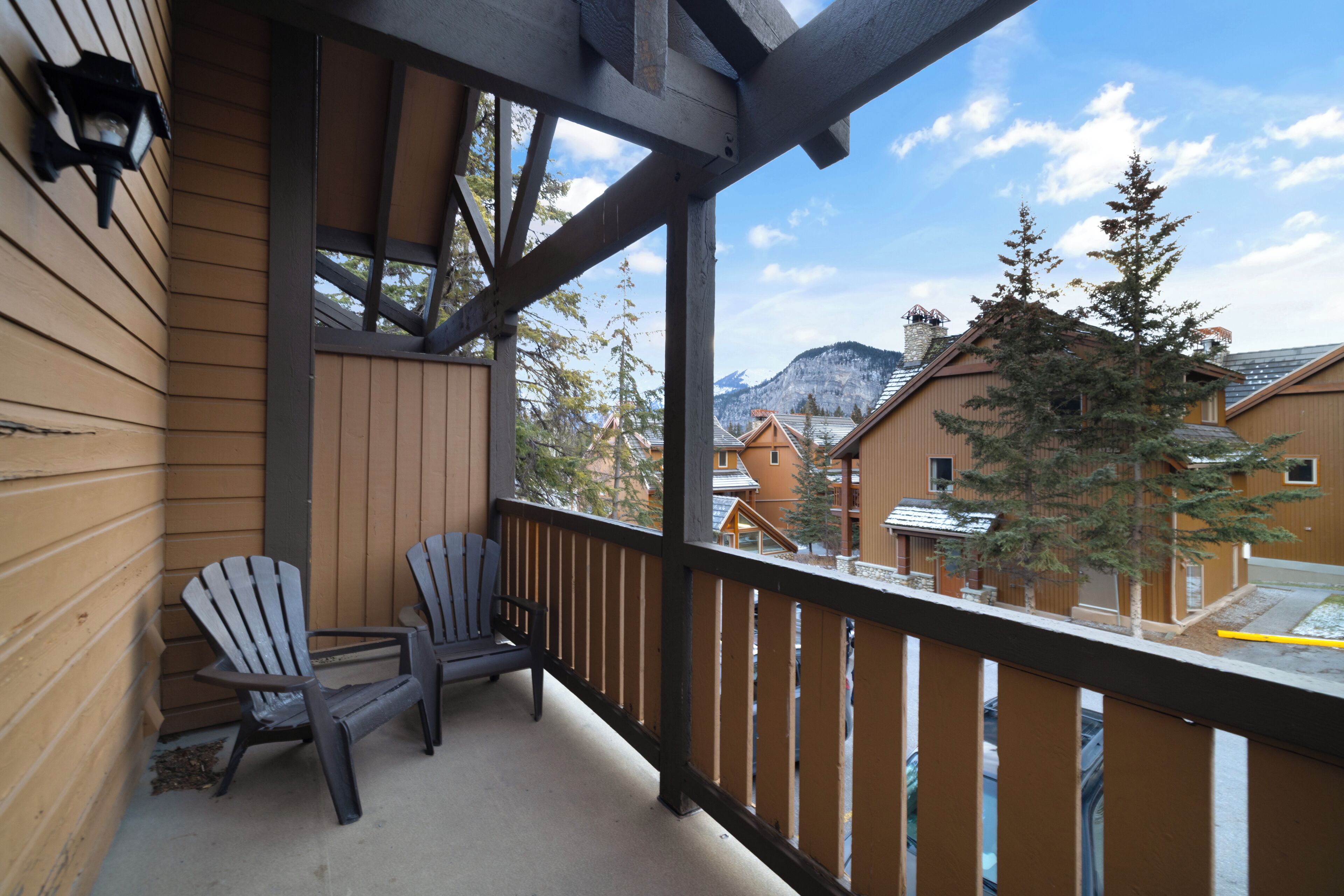 A wooden balcony with two Adirondack chairs, a view of a mountain and other buildings.