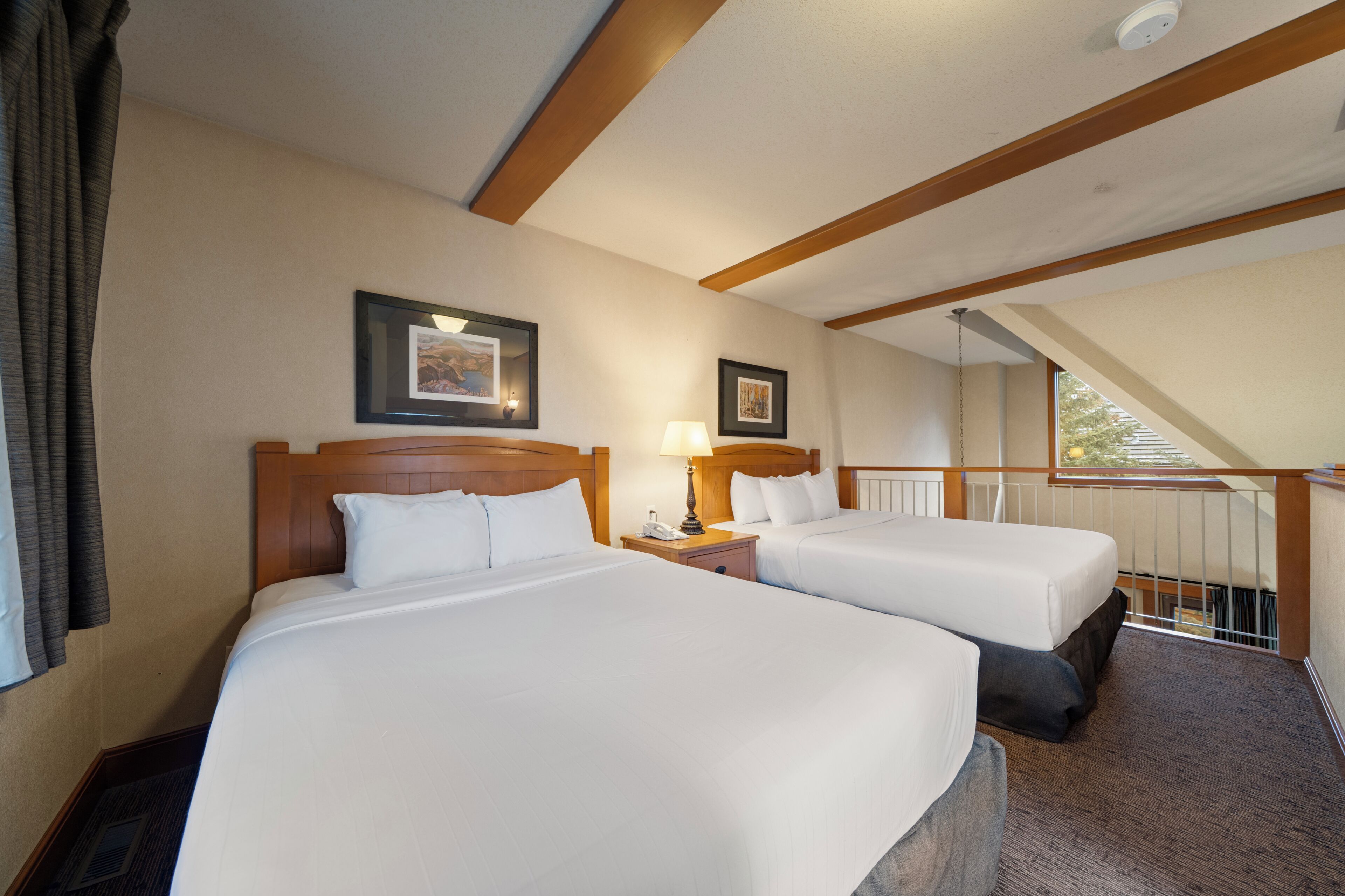 A hotel room with two beds, a nightstand with a lamp, a framed picture on the wall, and a window with a view of trees.