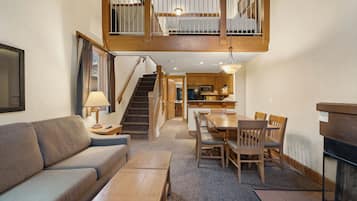 Una sala de estar acogedora con un sofá, una mesa de comedor de madera y sillas. Una escalera conduce a un nivel superior con un balcón.