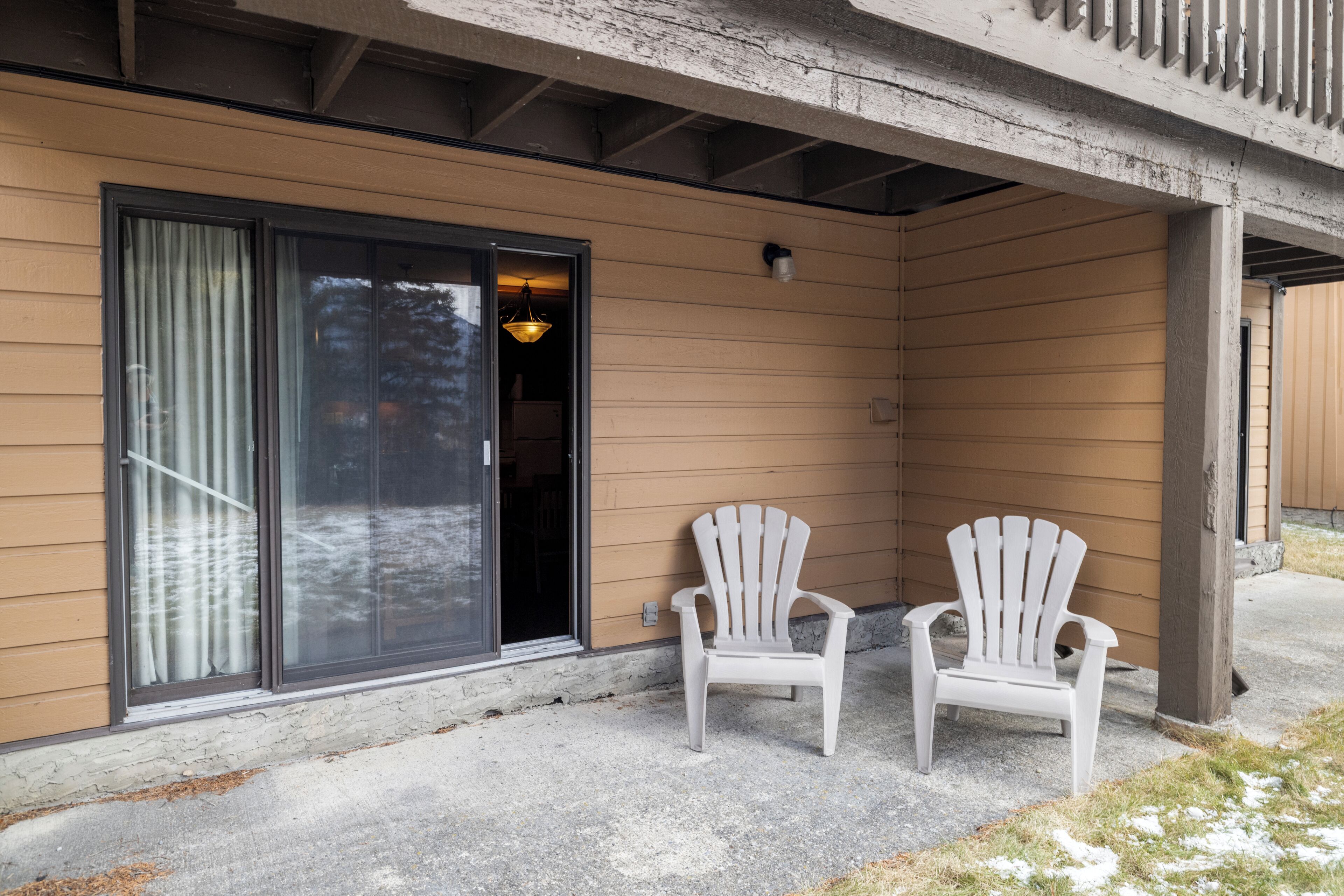 A building with a covered porch, two white Adirondack chairs, and a sliding glass door.