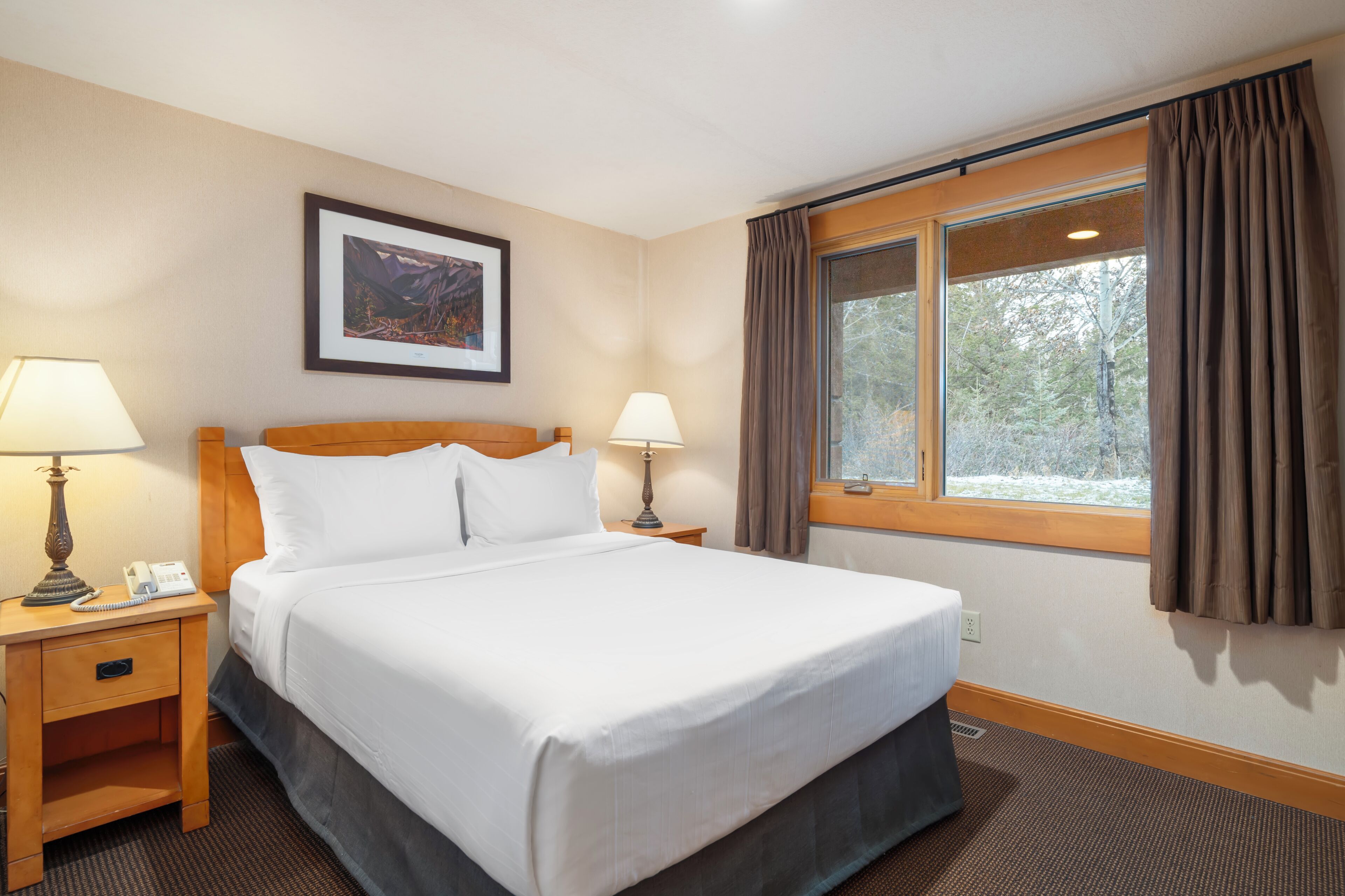 A hotel room with a bed, two bedside lamps, a telephone, a framed picture on the wall, and a window with curtains.