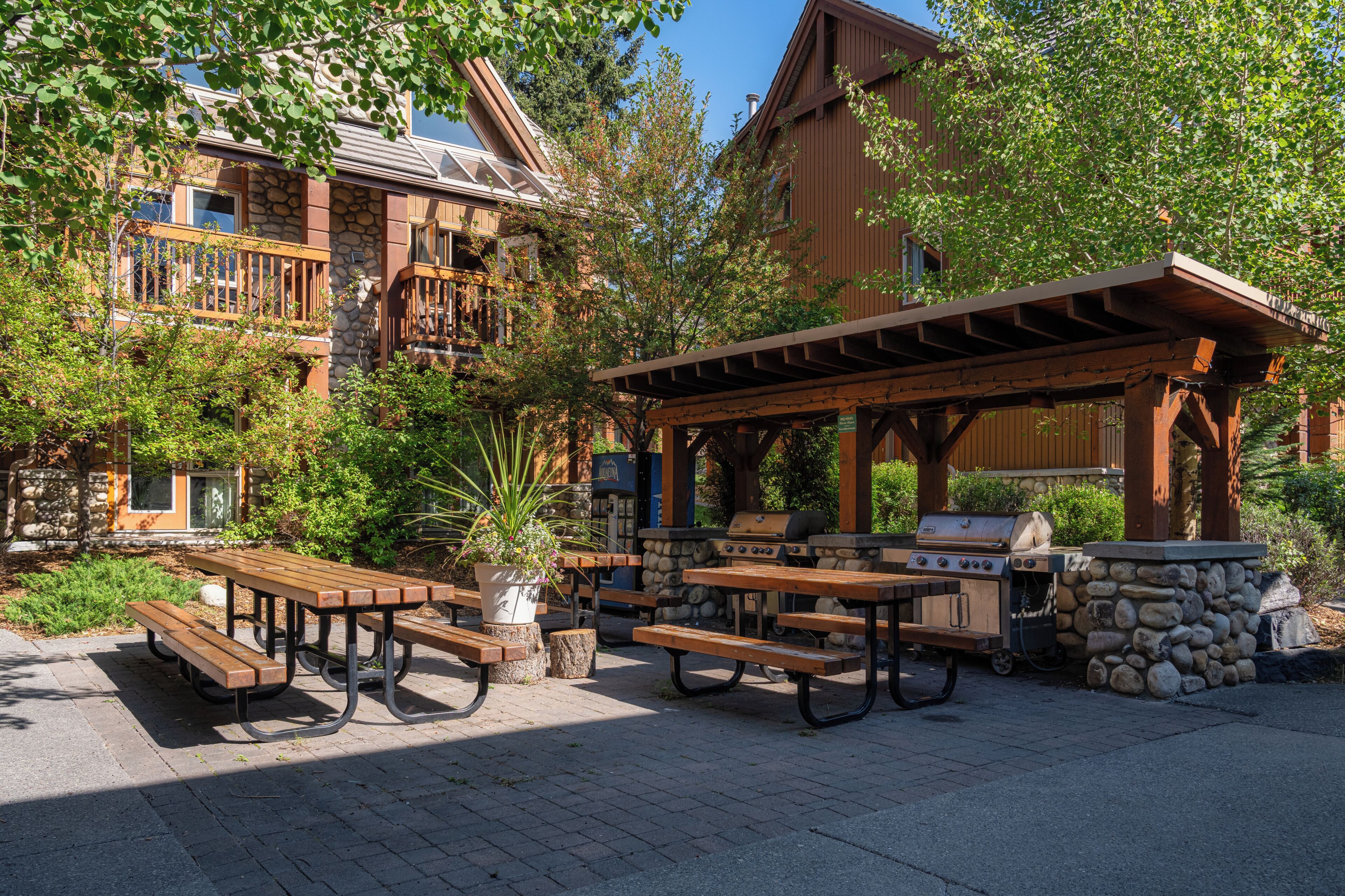 Un patio al aire libre con mesas de picnic y una parrilla, rodeado de muros de piedra y estructuras de madera.