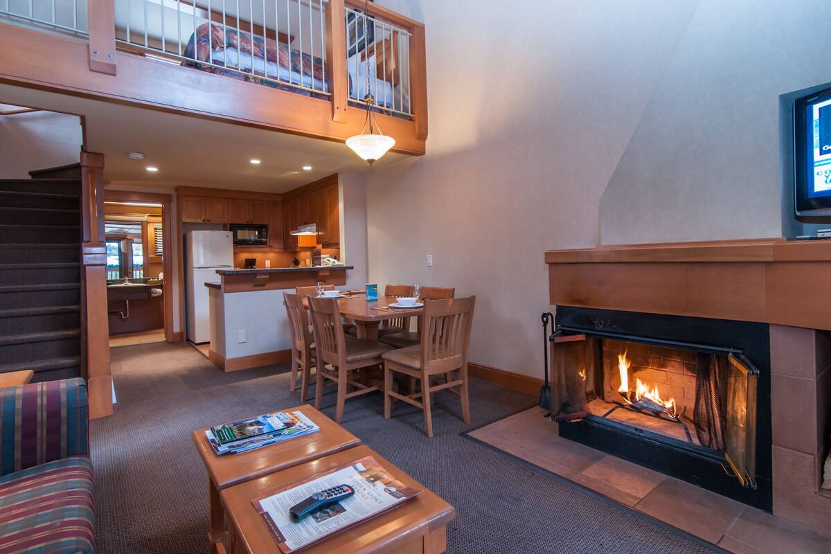 A spacious living area with a fireplace, a dining table with chairs, a kitchen in the background, and a balcony with a railing.