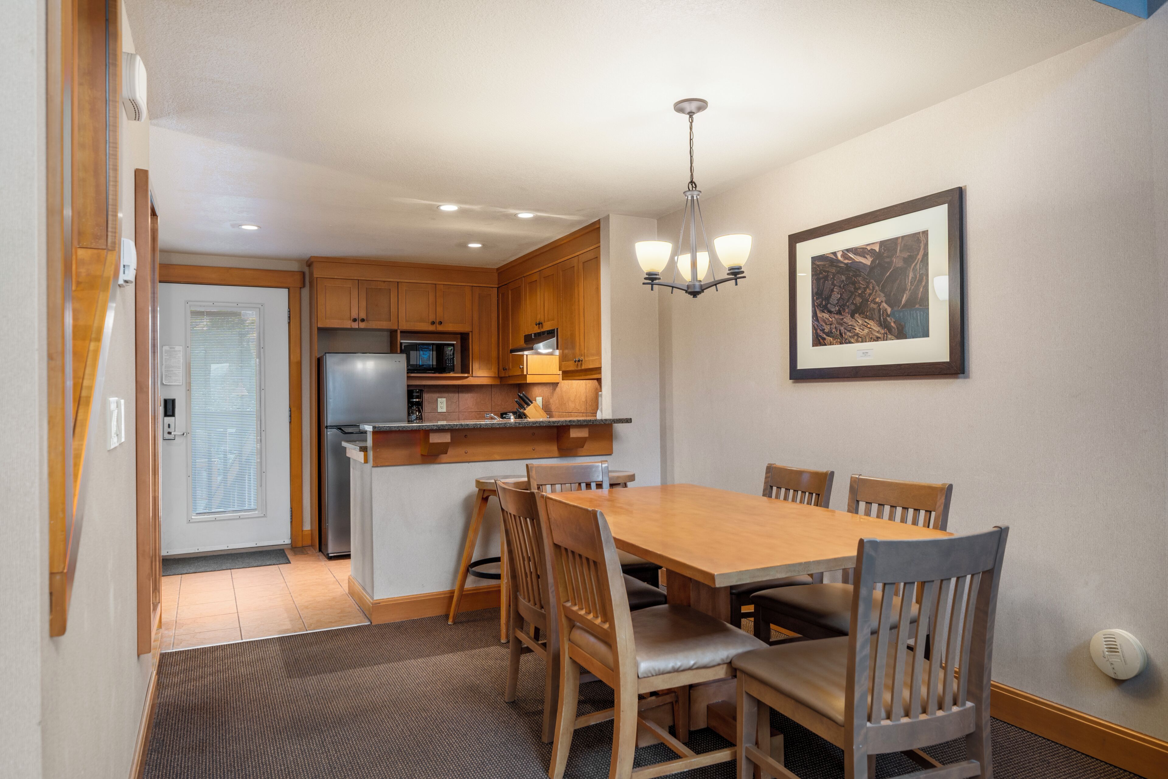 A dining area with a wooden table and chairs, a kitchen with wooden cabinets, a refrigerator, and a microwave. There is a framed picture on the wall and a ceiling light fixture.