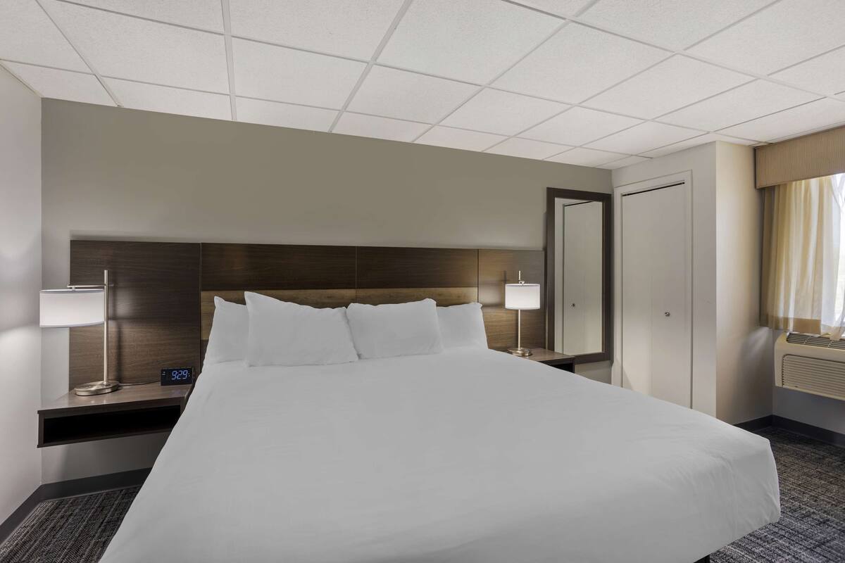 A hotel room with a large bed, bedside tables with lamps, a digital clock, a mirror, and a window with curtains.