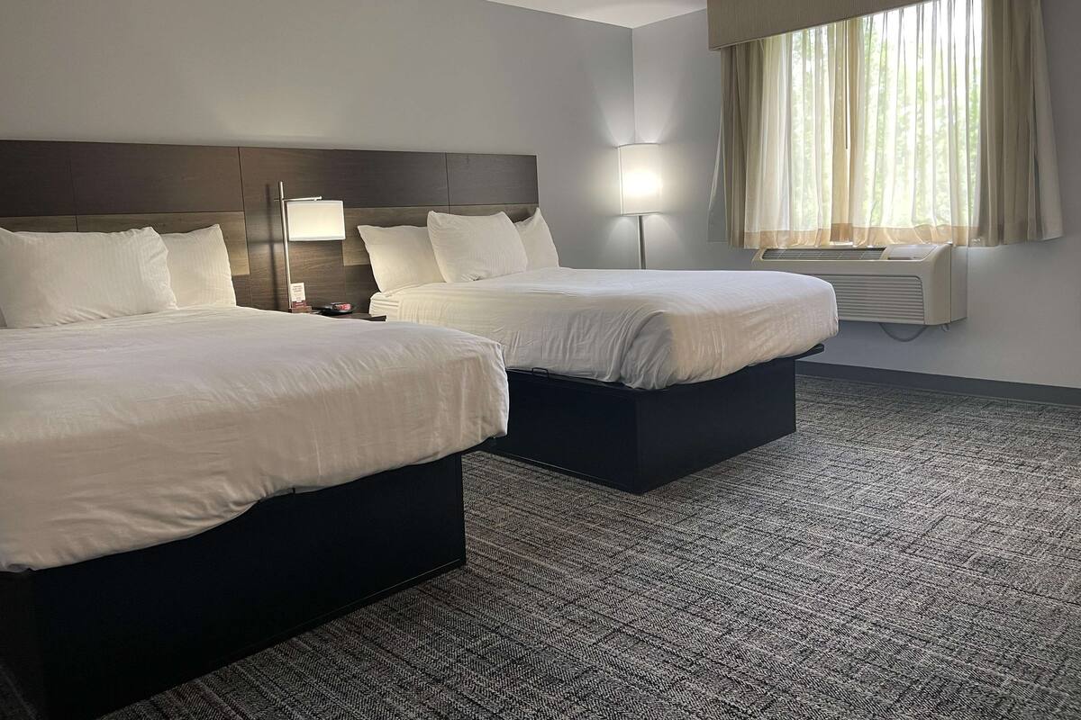 A hotel room with two beds, a headboard, a bedside table, a lamp, an air conditioner, and a window with curtains.