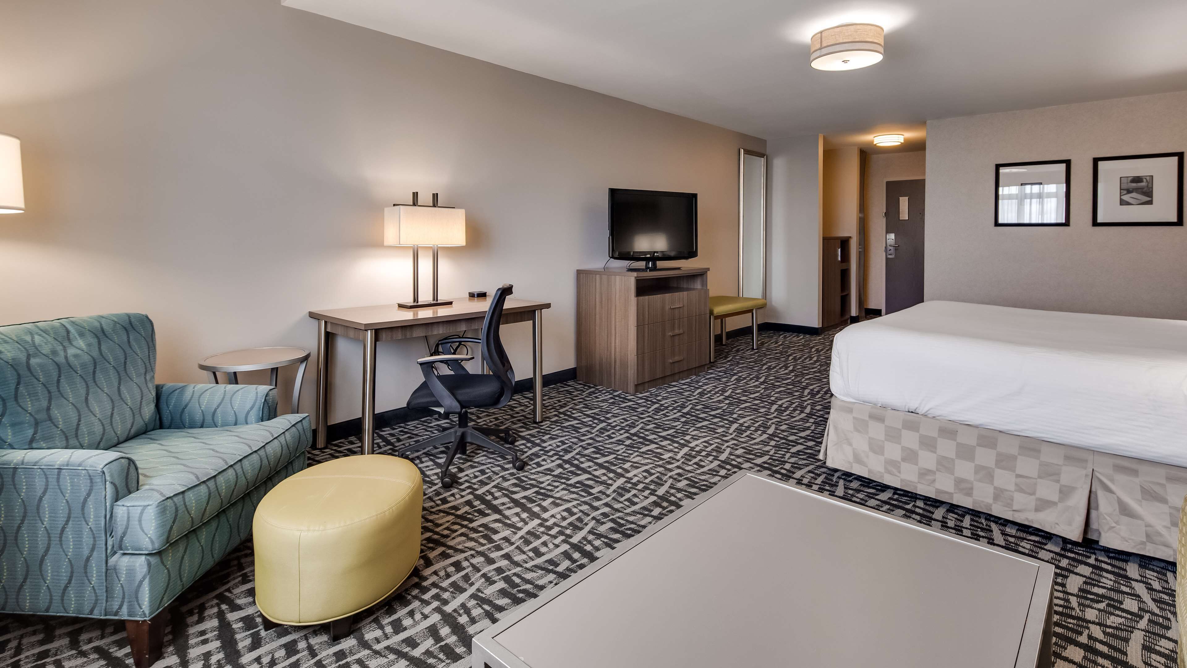 A hotel room with a bed, a desk with a chair, a sofa, a small table, a lamp, and a television.