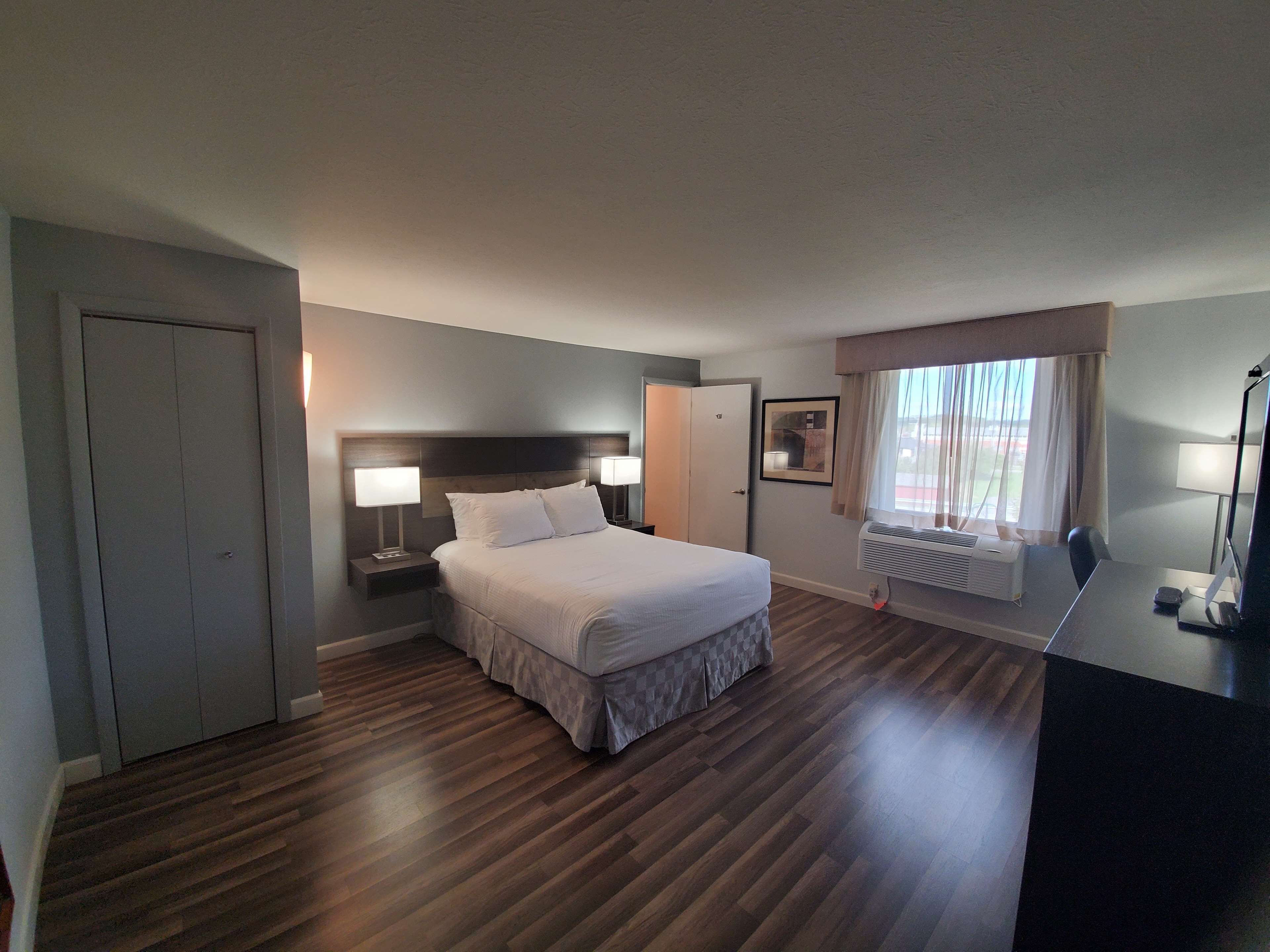 A hotel room with a bed, bedside lamps, a desk with a computer, a window with curtains, and a radiator.