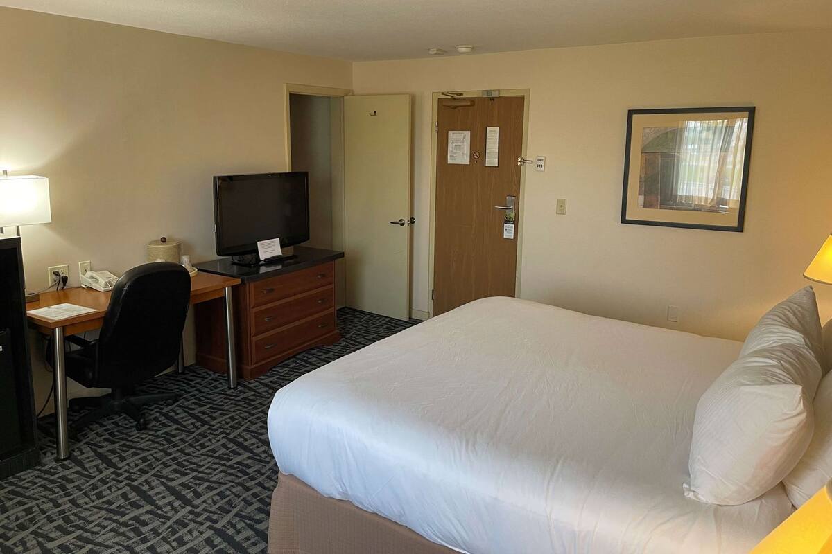 A hotel room with a bed, a desk with a computer and a television, a chair, and a door.