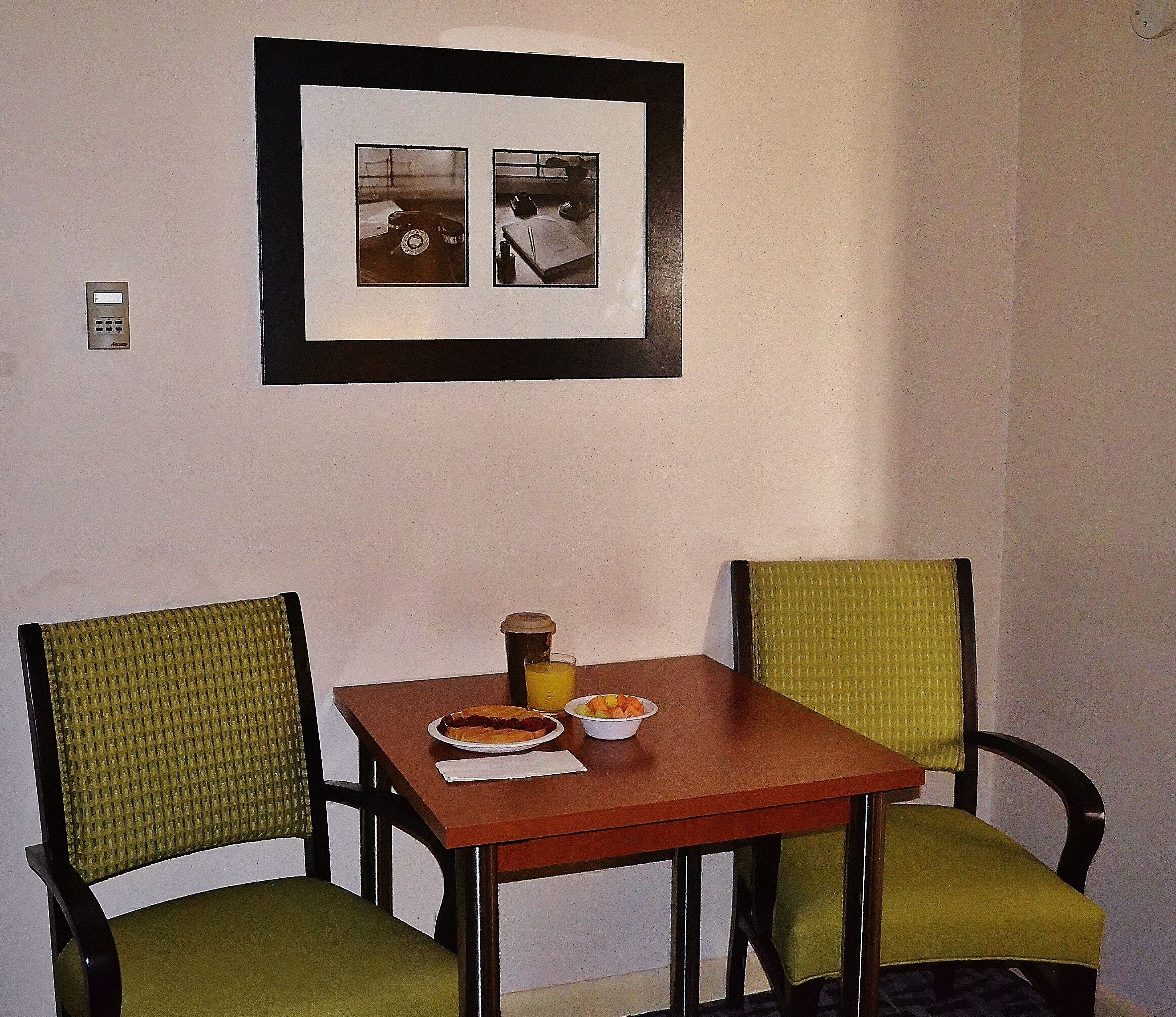 Un pequeño comedor con una mesa y dos sillas, un cuadro enmarcado sobre la mesa y un interruptor de luz en la pared.