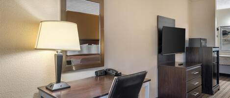 A hotel room with a desk, chair, lamp, mirror, television, and a wardrobe.