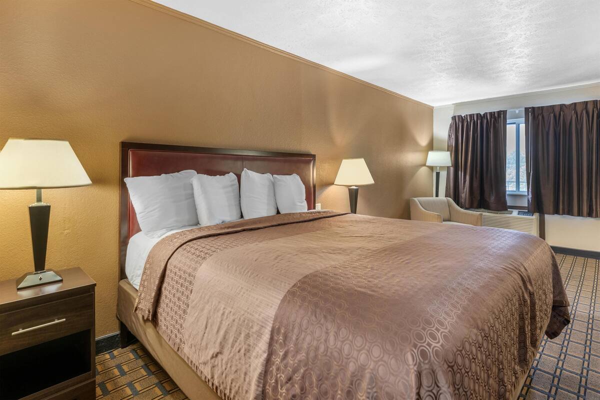 Habitación de hotel con una cama grande, dos mesitas de noche con lámparas, una silla y un ventanal con cortinas.