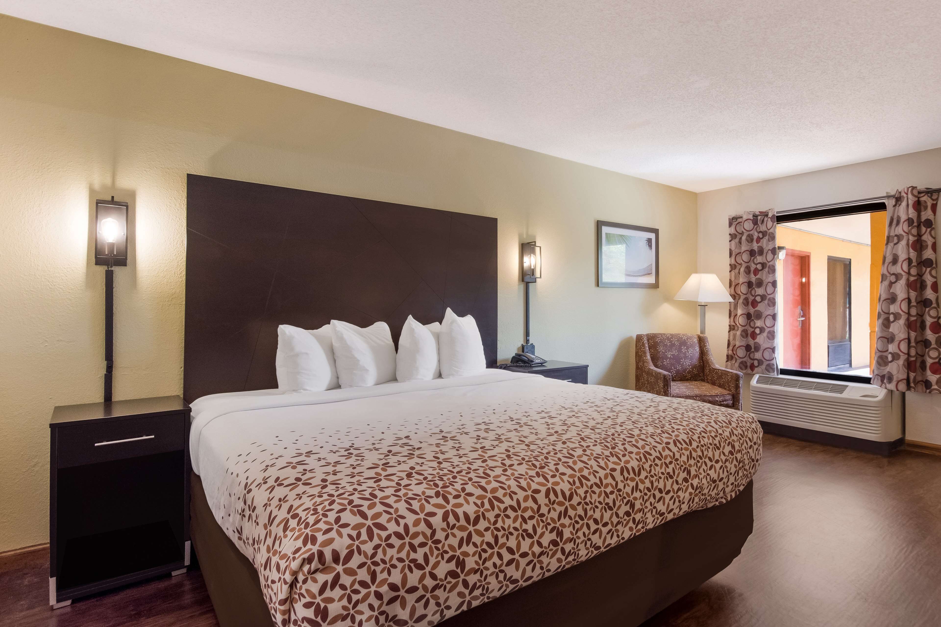 A hotel room with a large bed, a nightstand, a chair, a lamp, and a window with curtains.