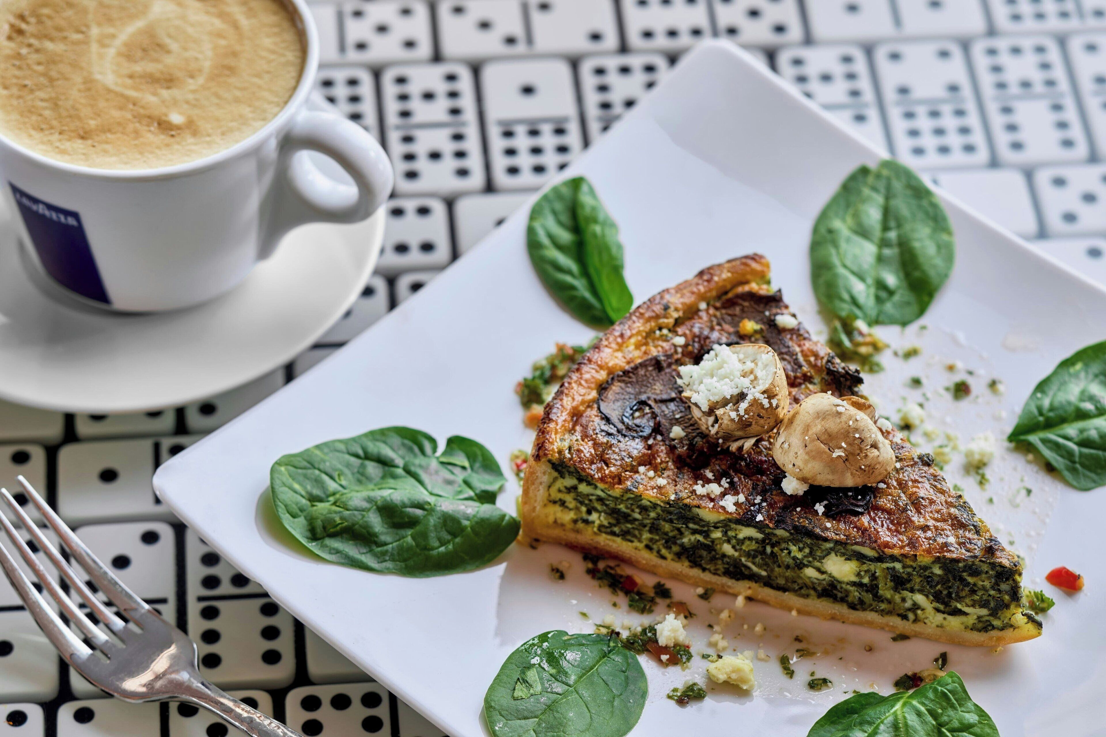 A slice of quiche on a white plate with spinach leaves and mushrooms, accompanied by a cup of coffee on a patterned table.