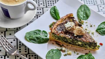 A slice of quiche on a white plate with spinach leaves and mushrooms, accompanied by a cup of coffee on a patterned table.