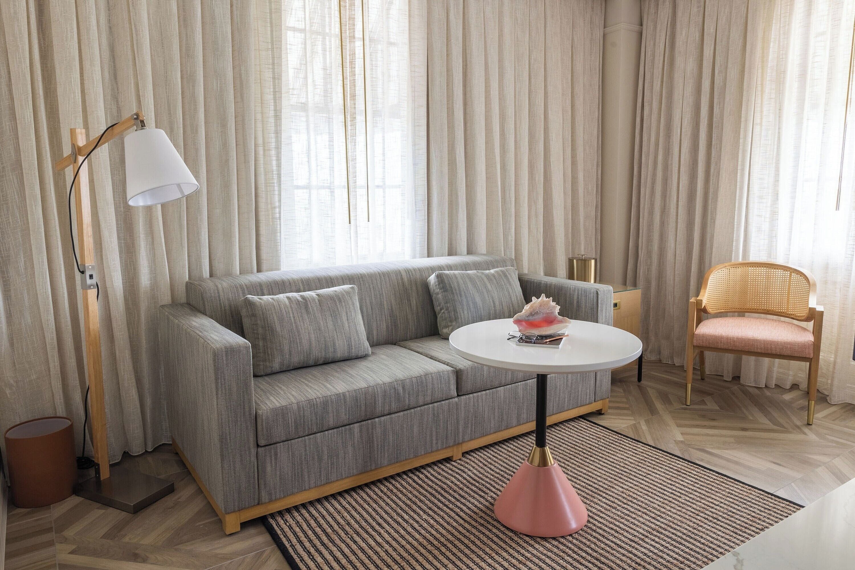 A modern living room with a grey sofa, a round white table, a pink floor lamp, and a wicker chair.