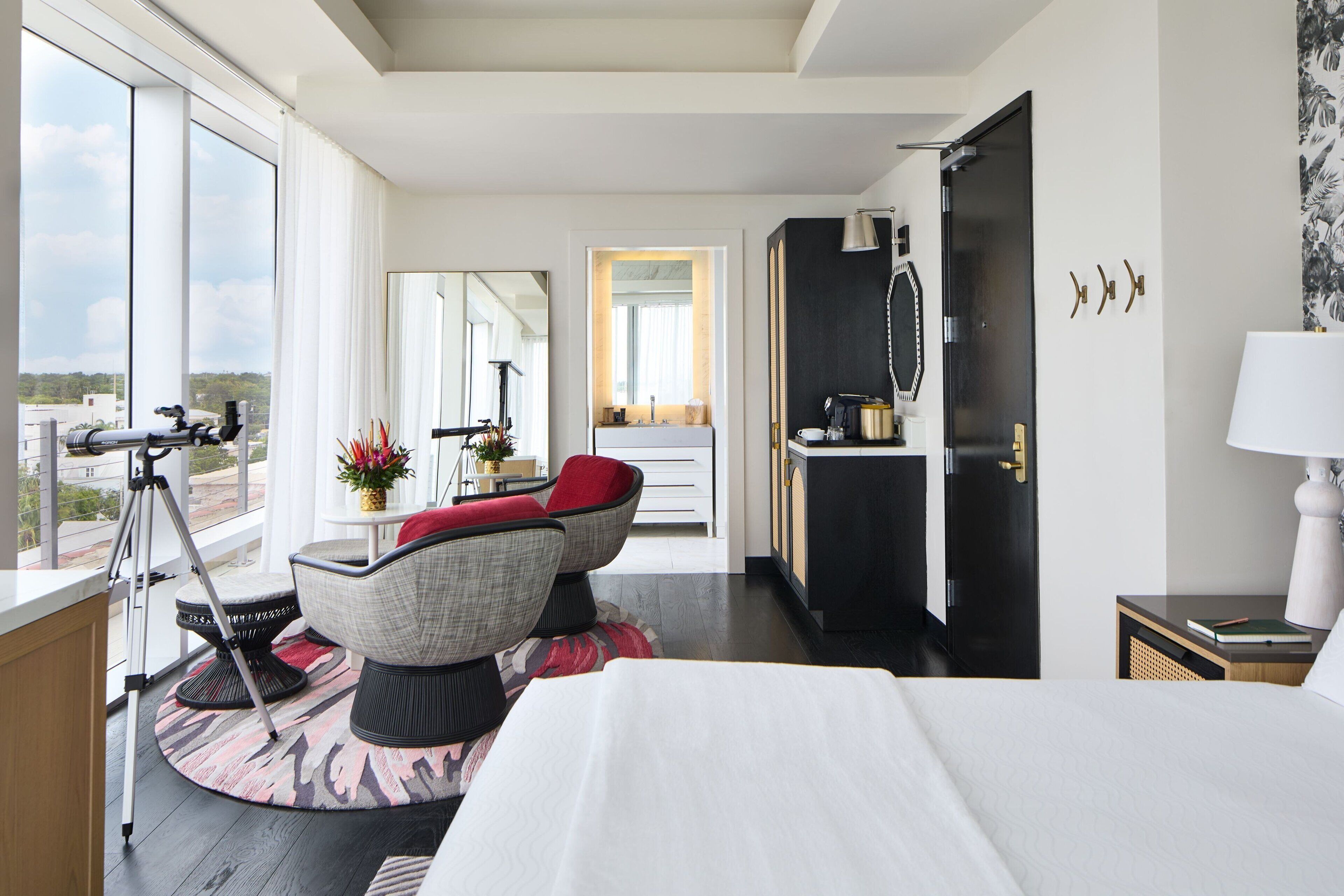 Une chambre d’hôtel moderne avec un grand lit, un coin salon avec un fauteuil rouge, un bureau avec une lampe et une vue sur la ville grâce à de grandes fenêtres.