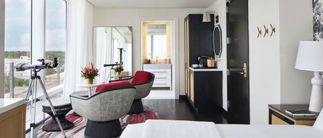 Ein modernes Hotelzimmer mit einem großen Bett, einer Sitzecke mit einem roten Sessel, einem Schreibtisch mit einer Lampe und Blick auf die Stadt durch große Fenster.