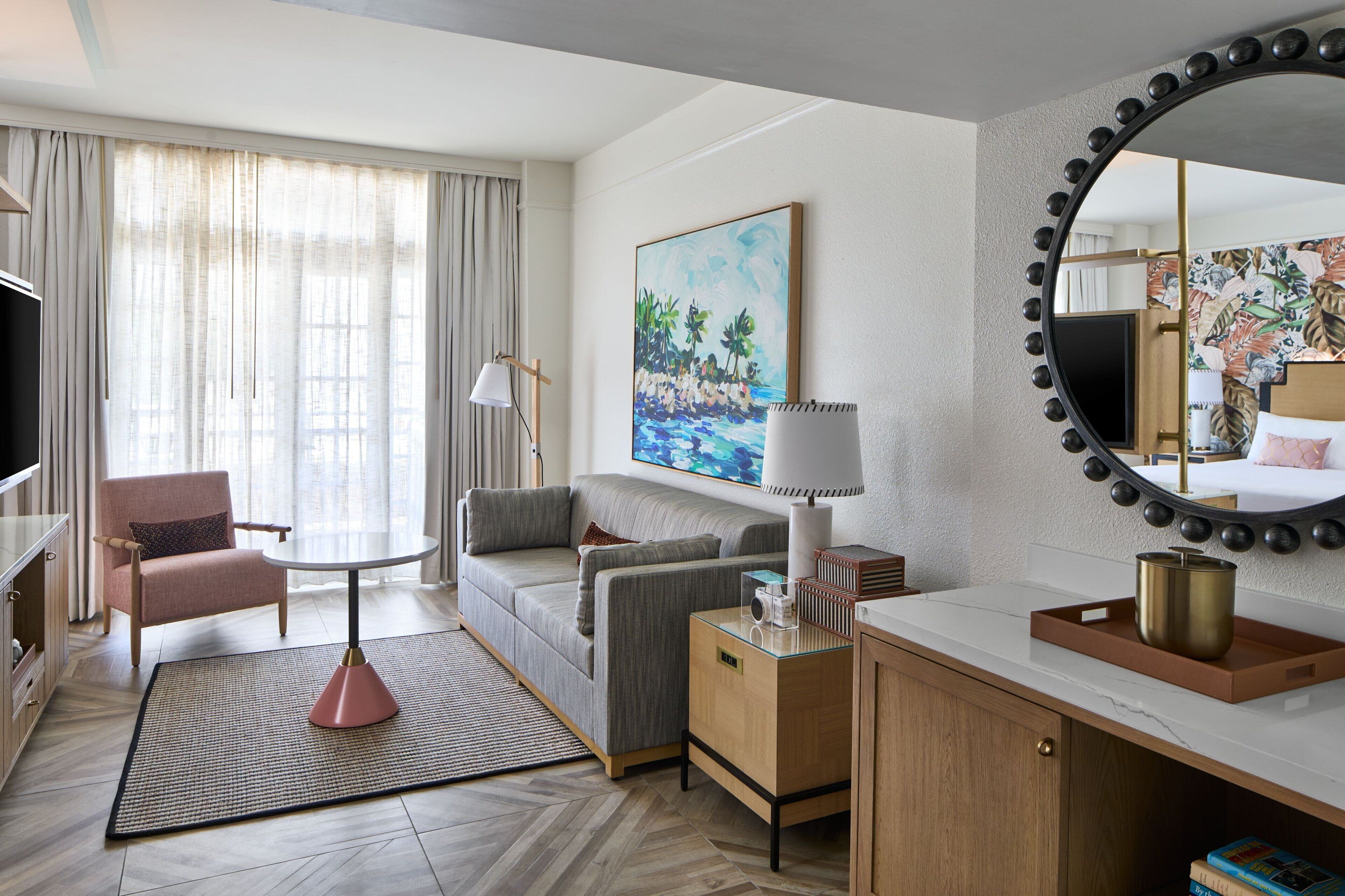 A hotel room with a sofa, armchair, TV, lamp, painting, and a large mirror.
