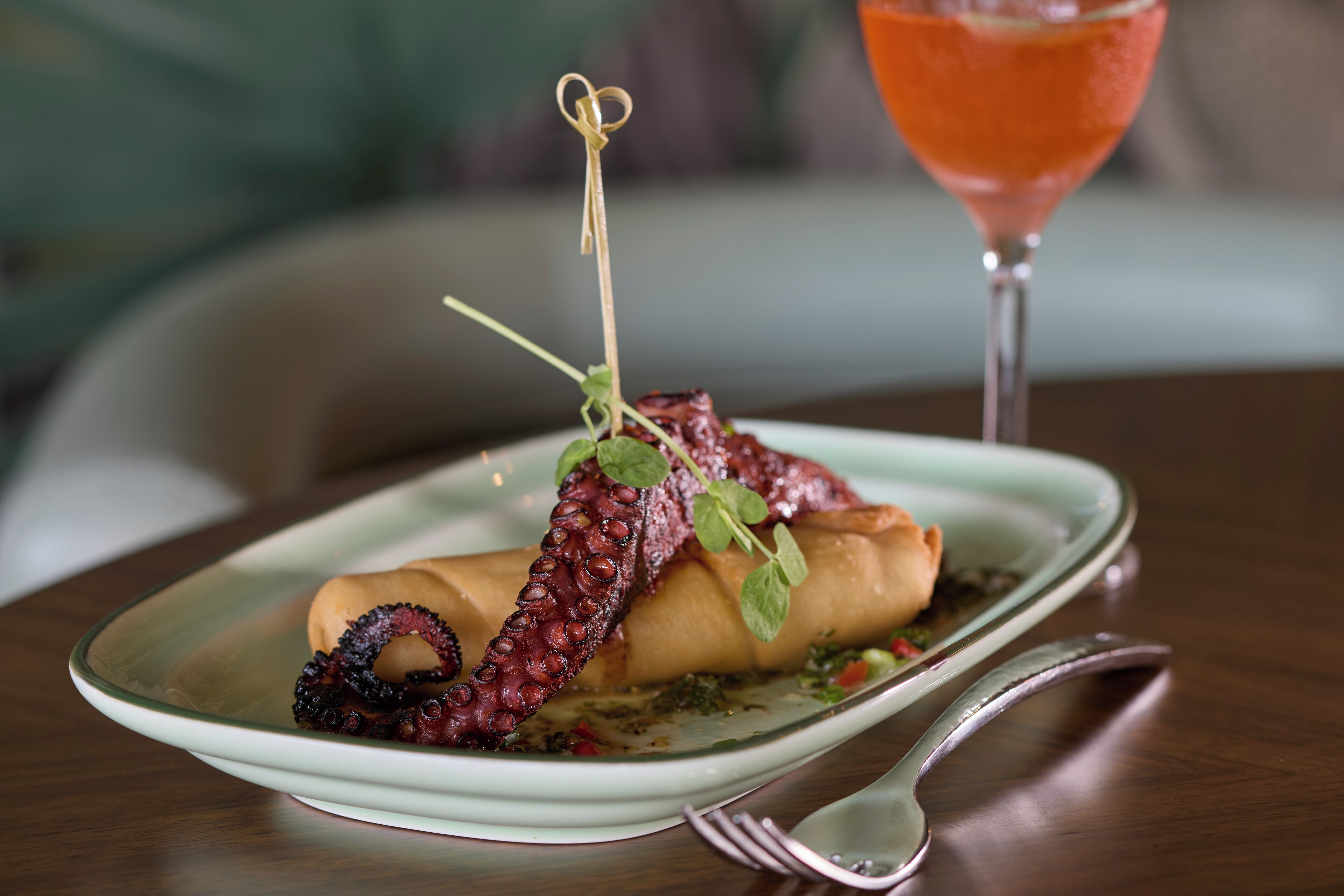 A plate with a rolled appetizer garnished with a tentacle, a cocktail glass, and a fork on a table.