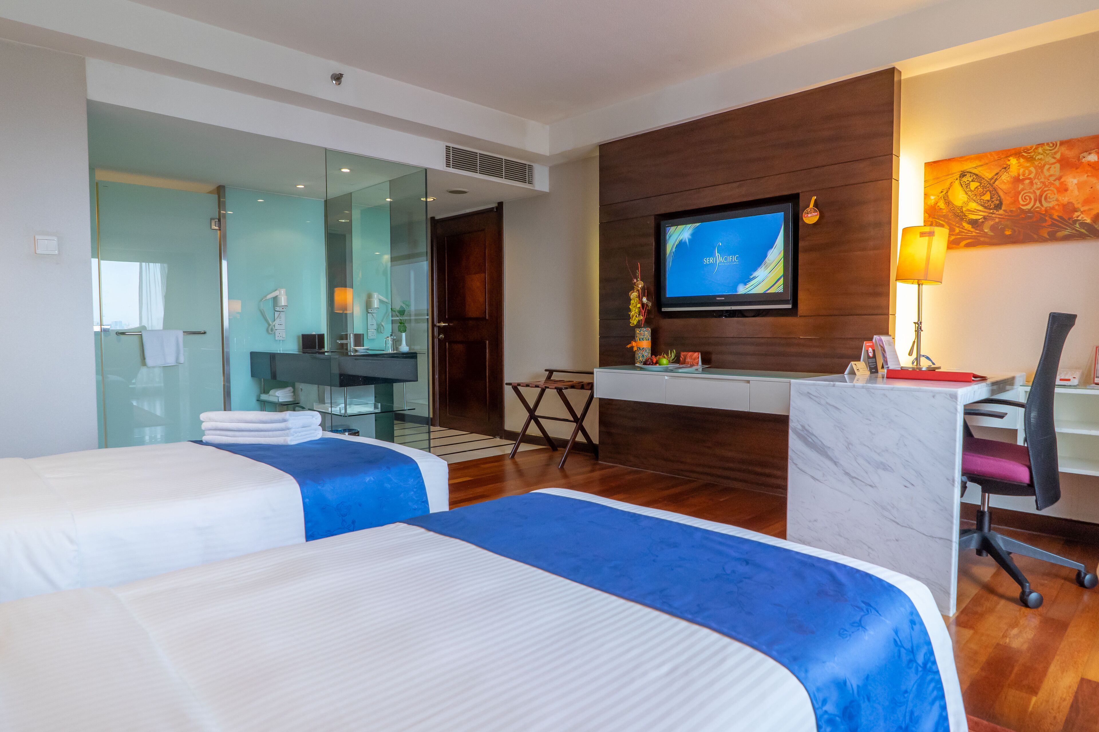 Habitación de hotel con dos camas, un escritorio, televisión y baño visible a través de una puerta de cristal.