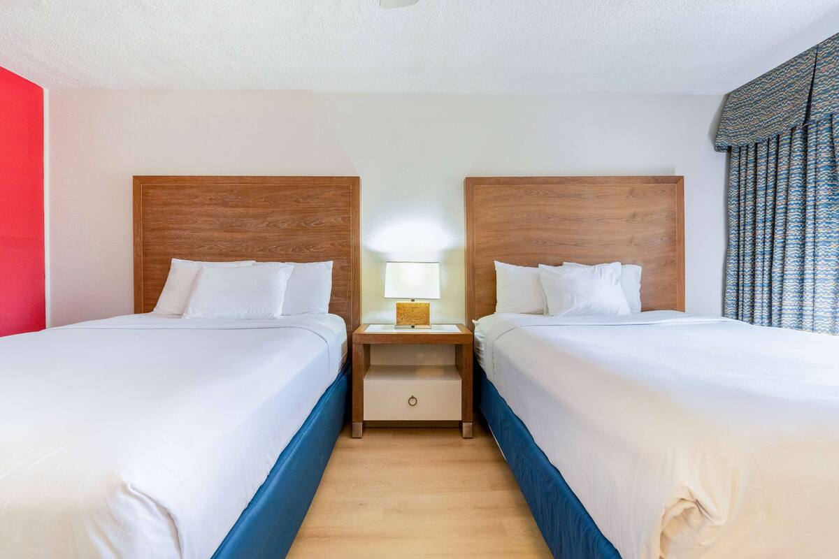 Een hotelkamer met twee eenpersoonsbedden, houten hoofdborden, witte beddengoed, een nachtkastje met een lamp en een raam met behang.