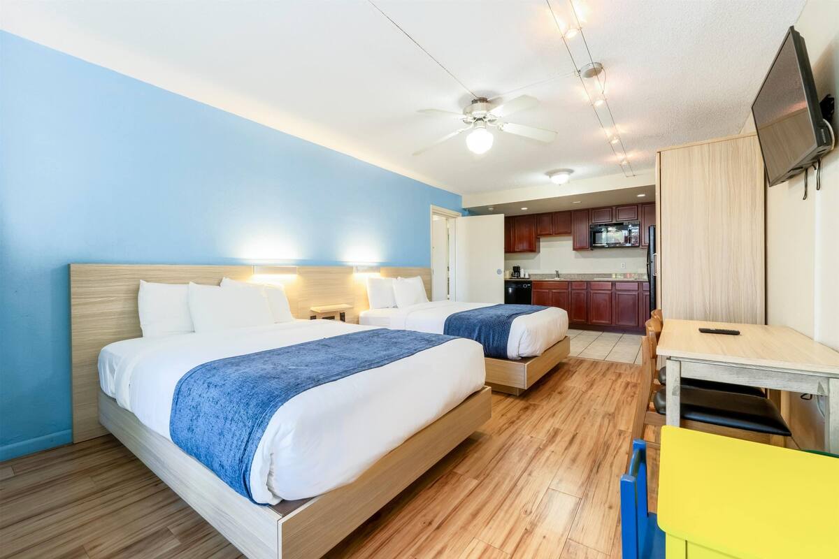 Hotelkamer met twee bedden, een bureau, een stoel, een televisie en een kitchenette op de achtergrond.