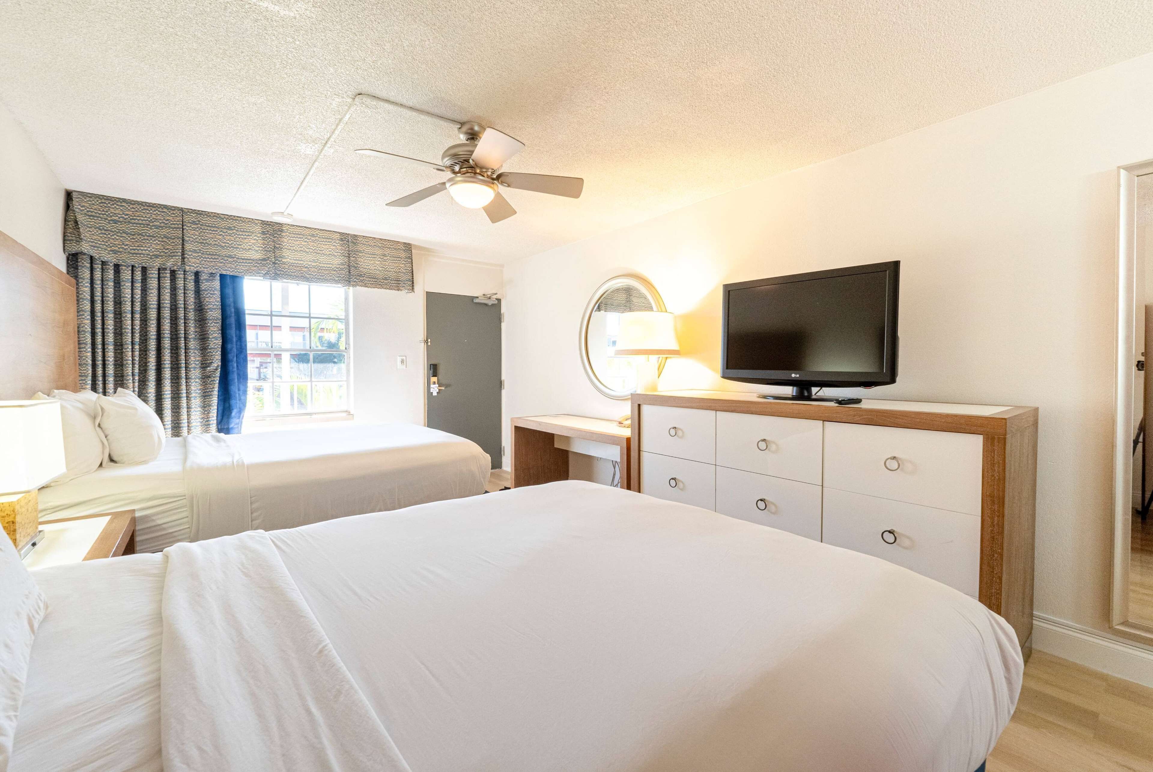 A hotel room with two beds, a desk with a lamp, a television, and a ceiling fan.