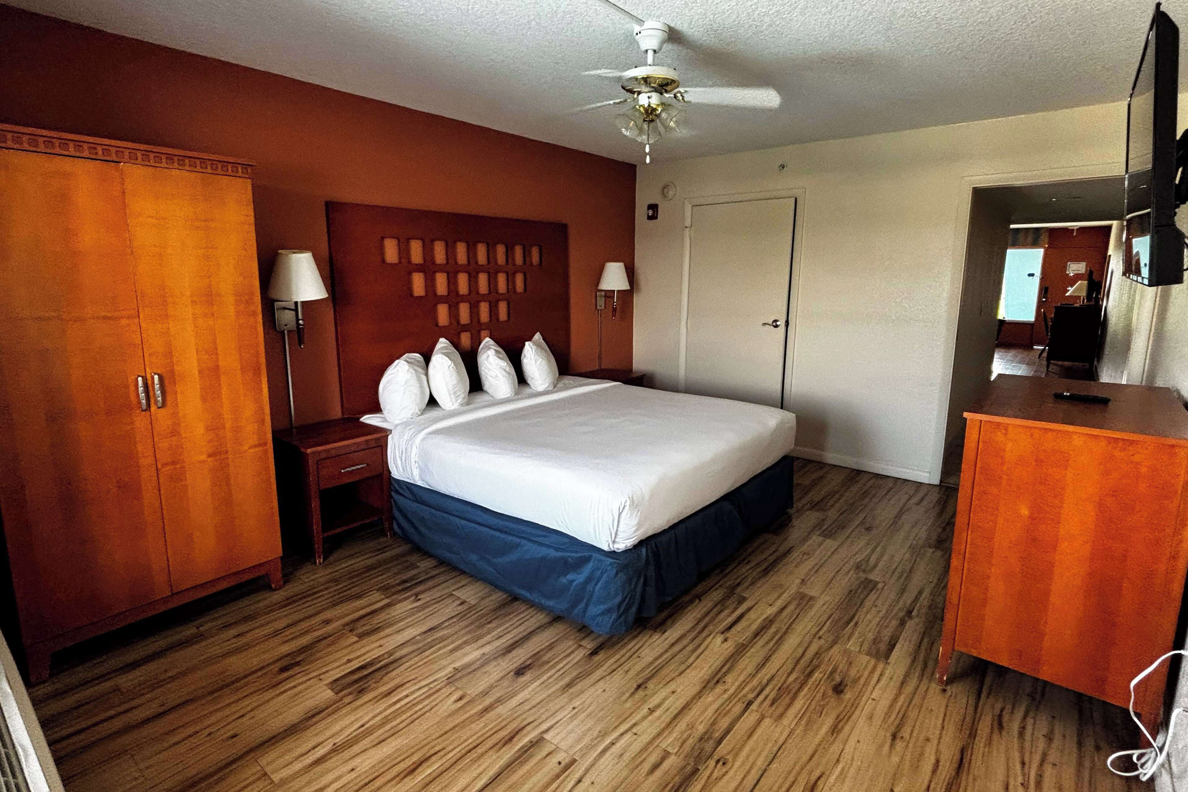 A hotel room with a wooden floor, a bed with a blue skirt, a wooden wardrobe, a nightstand, two wall-mounted lamps, a ceiling fan, and a television on a wooden cabinet.