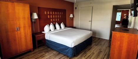 A hotel room with a wooden floor, a bed with a blue skirt, a wooden wardrobe, a nightstand, two wall-mounted lamps, a ceiling fan, and a television on a wooden cabinet.