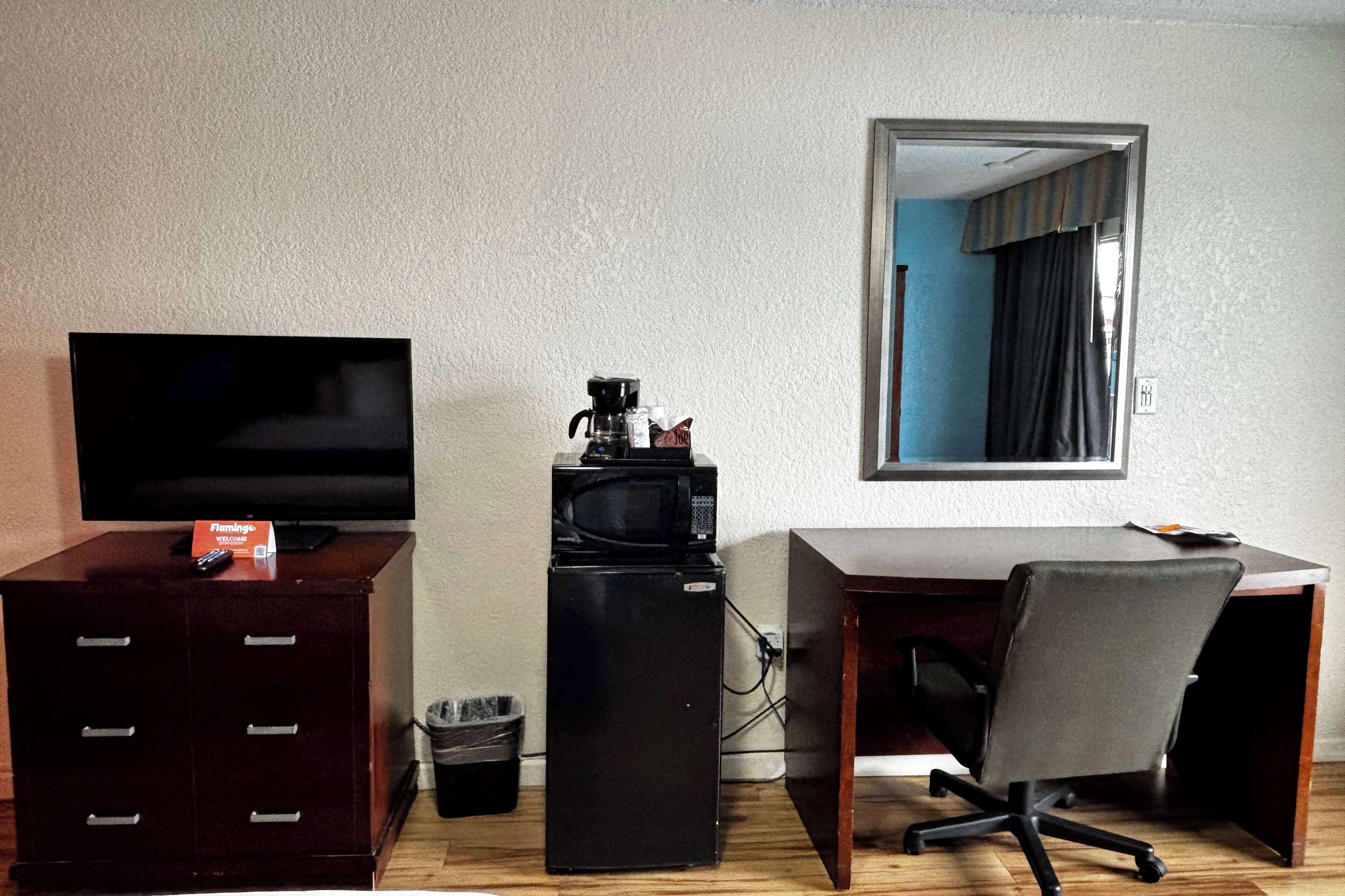A compact room with a flat-screen TV on a wooden cabinet, a small black refrigerator, a desk with a chair, and a mirror on the wall.