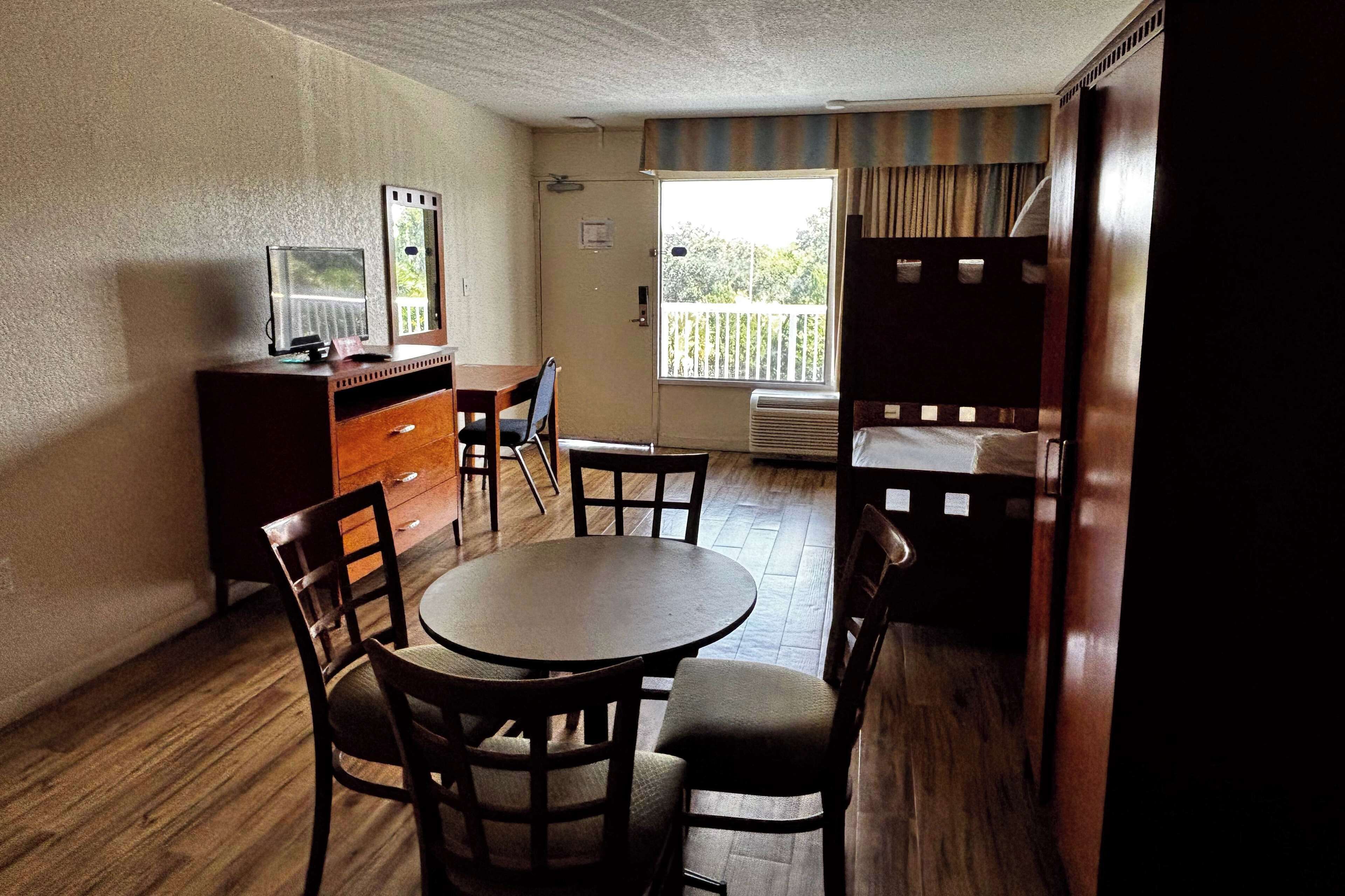 A compact room with a round table and four chairs, a small desk with a television, and a bunk bed against the wall.