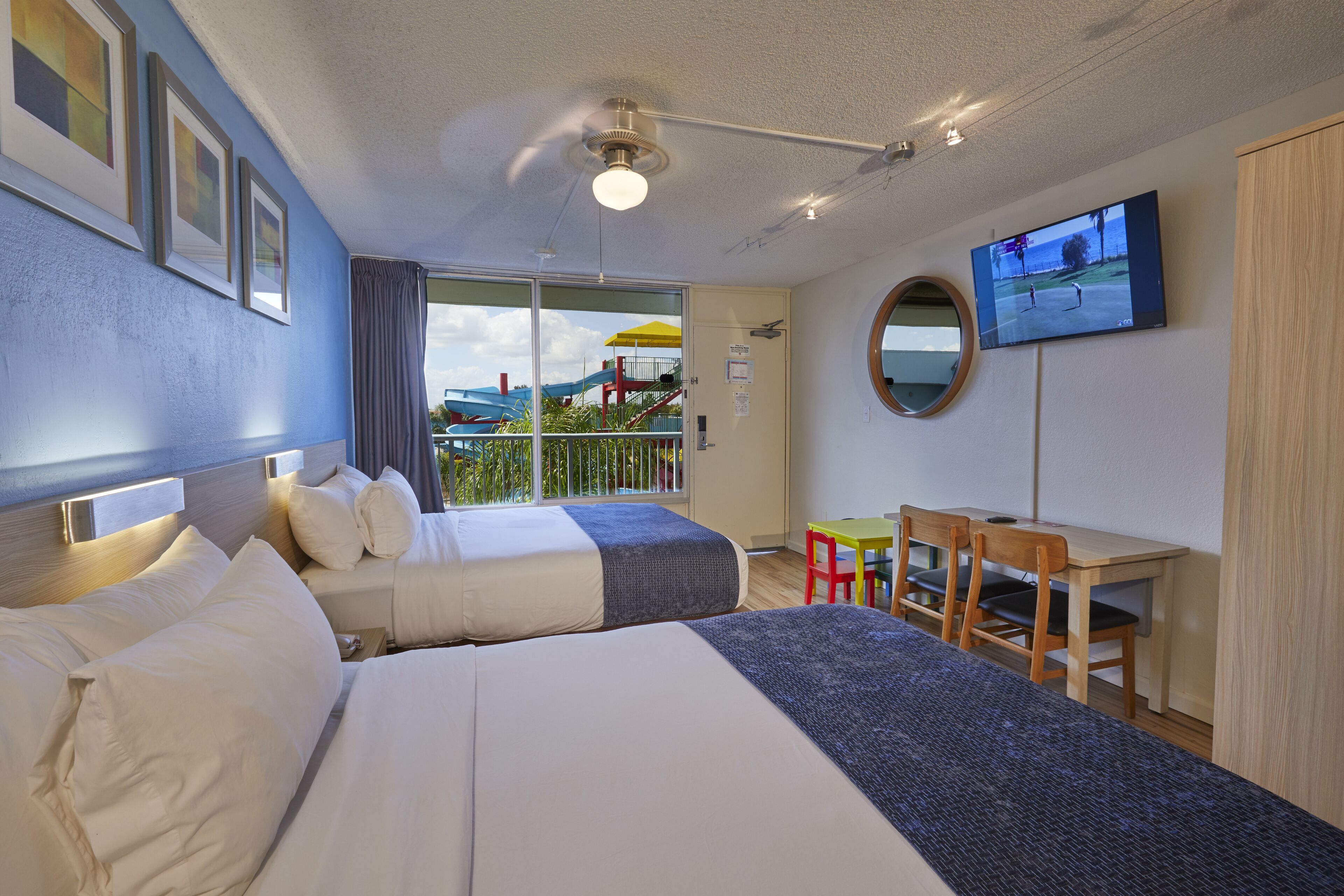 A hotel room with two beds, a desk, a chair, a TV, and a balcony with a view.