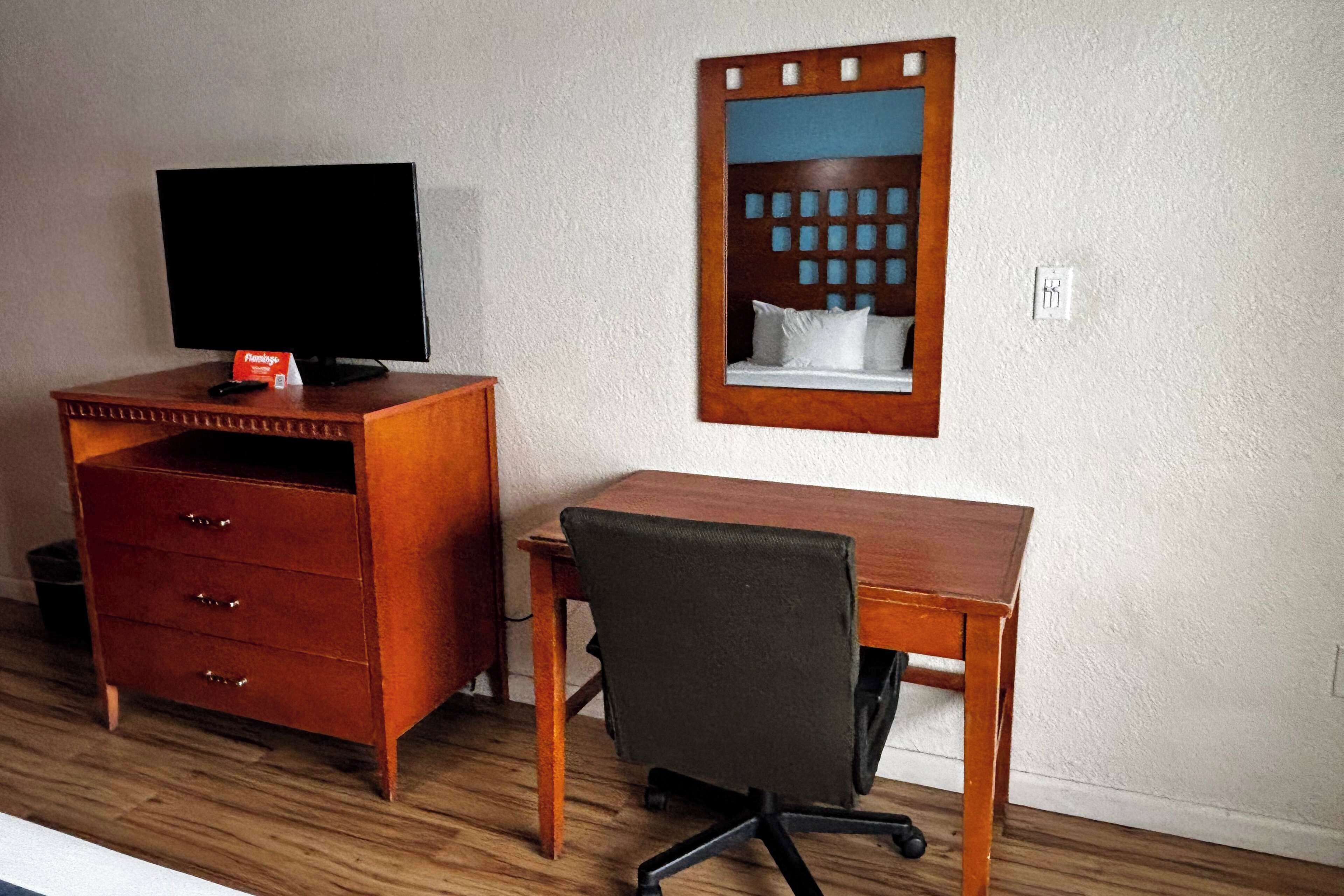 Una habitación con un escritorio de madera, una silla, un televisor sobre un aparador y un espejo en la pared.