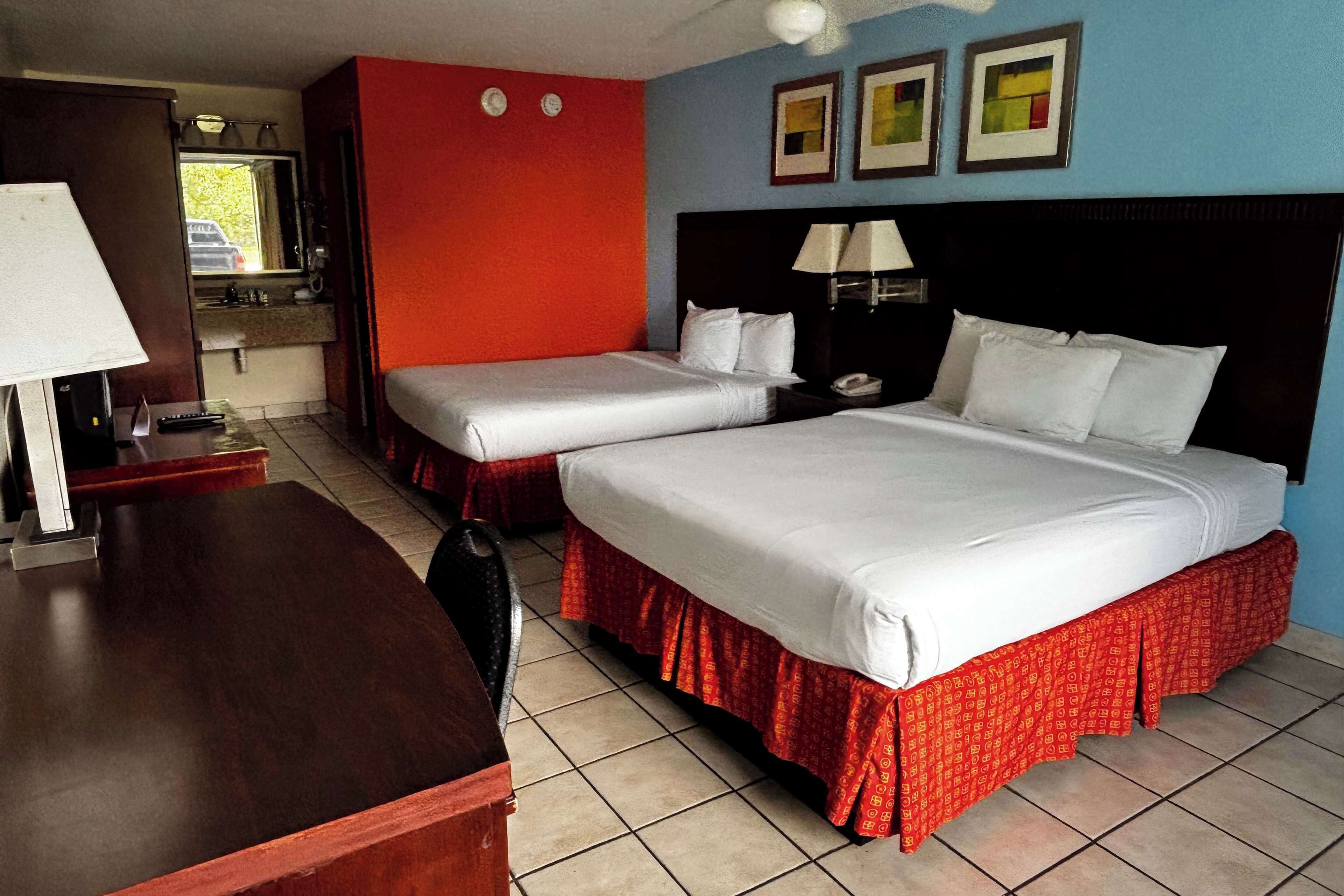 A hotel room with two beds, a desk, a chair, a mirror, a window, and a bathroom.