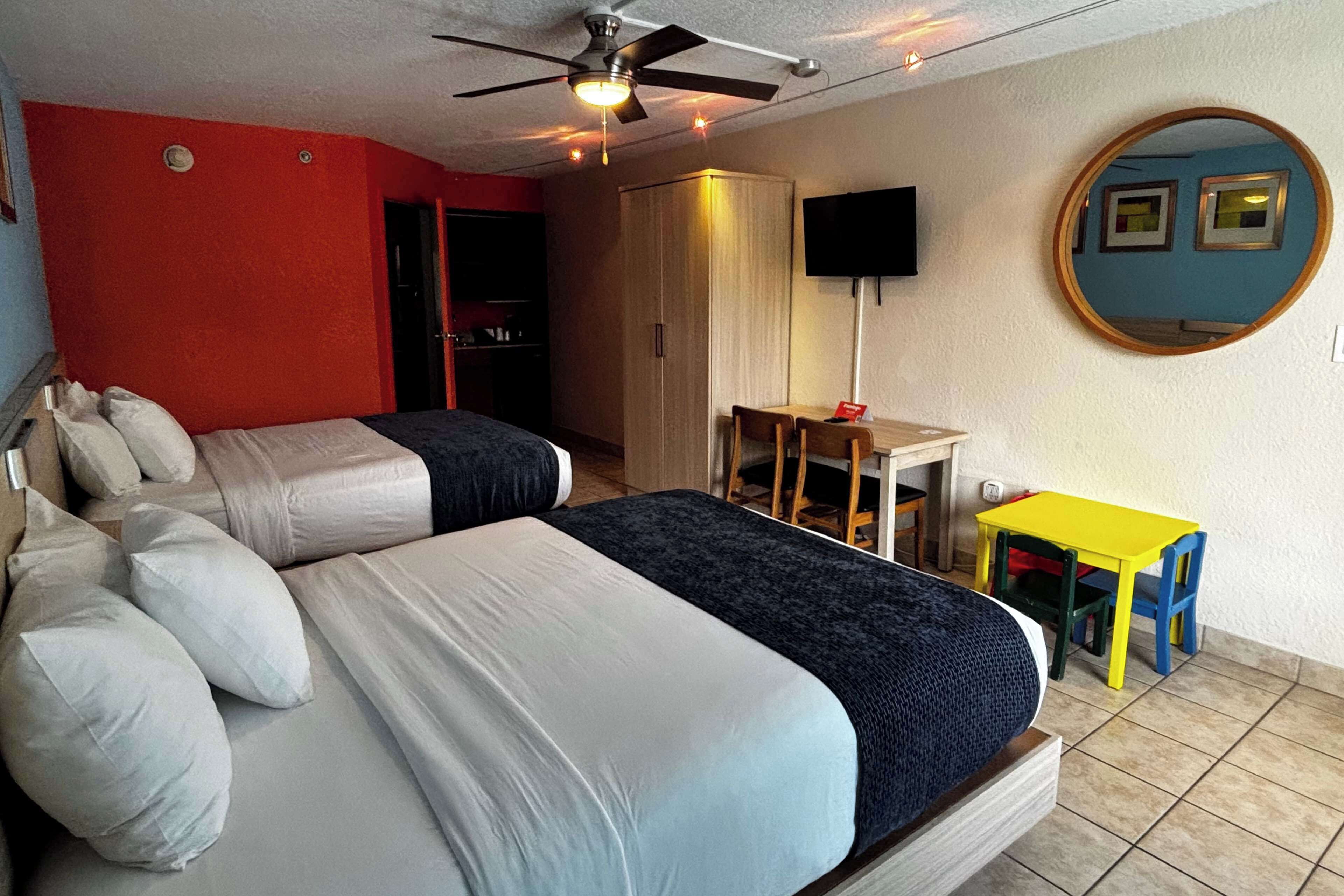 Camera d'albergo con due letti, una ventola a soffitto, una televisione, uno specchio e un piccolo tavolo con sedie.